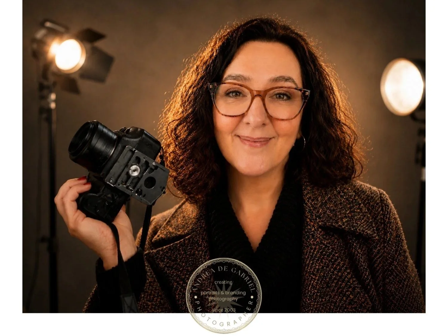A woman with curly dark hair and glasses holding a camera in a photography studio with professional lighting around her.