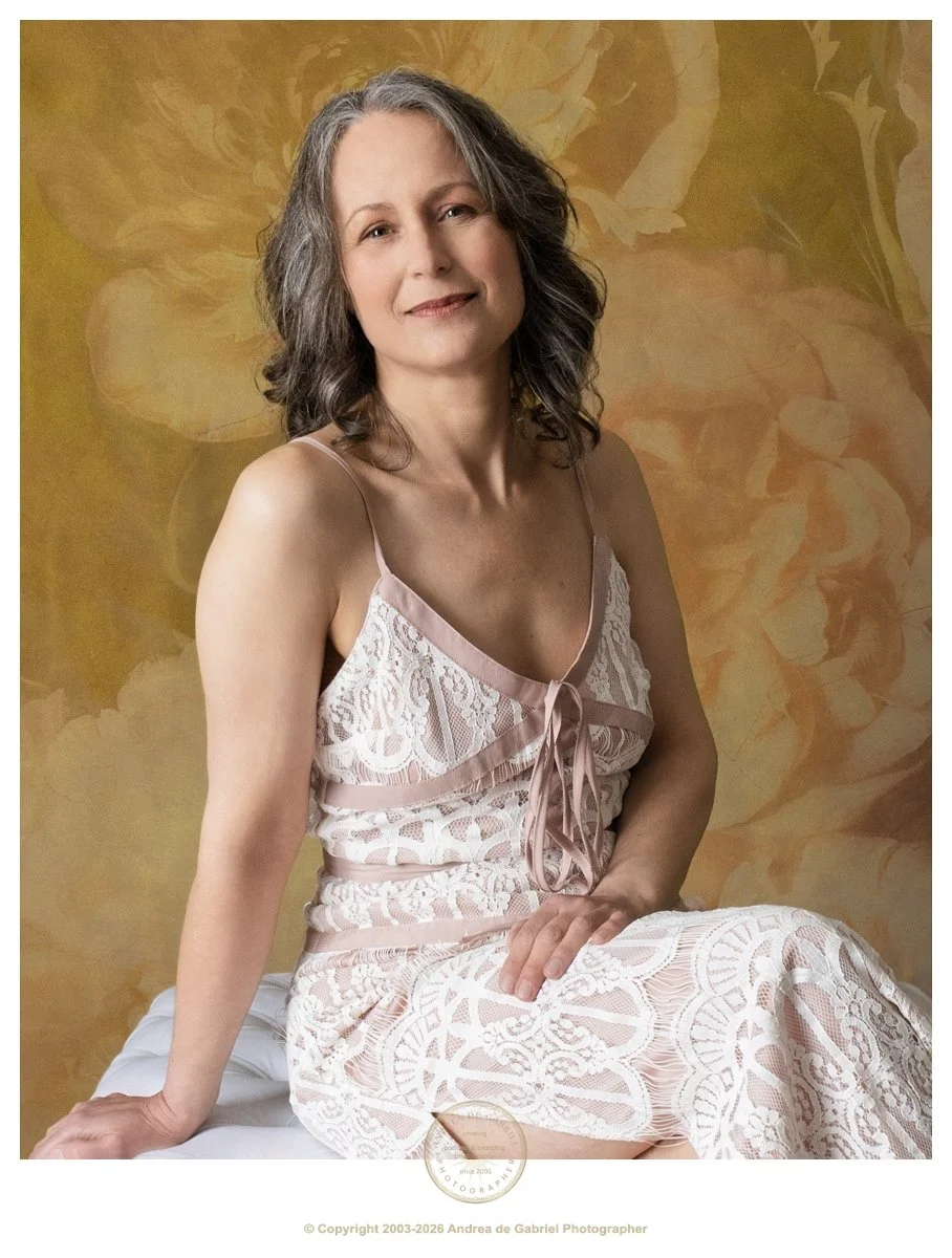 mature woman with shoulder length silver hair, sits with hand on her lap, looking at the camera with a seer expression, wearing a pink lace dress