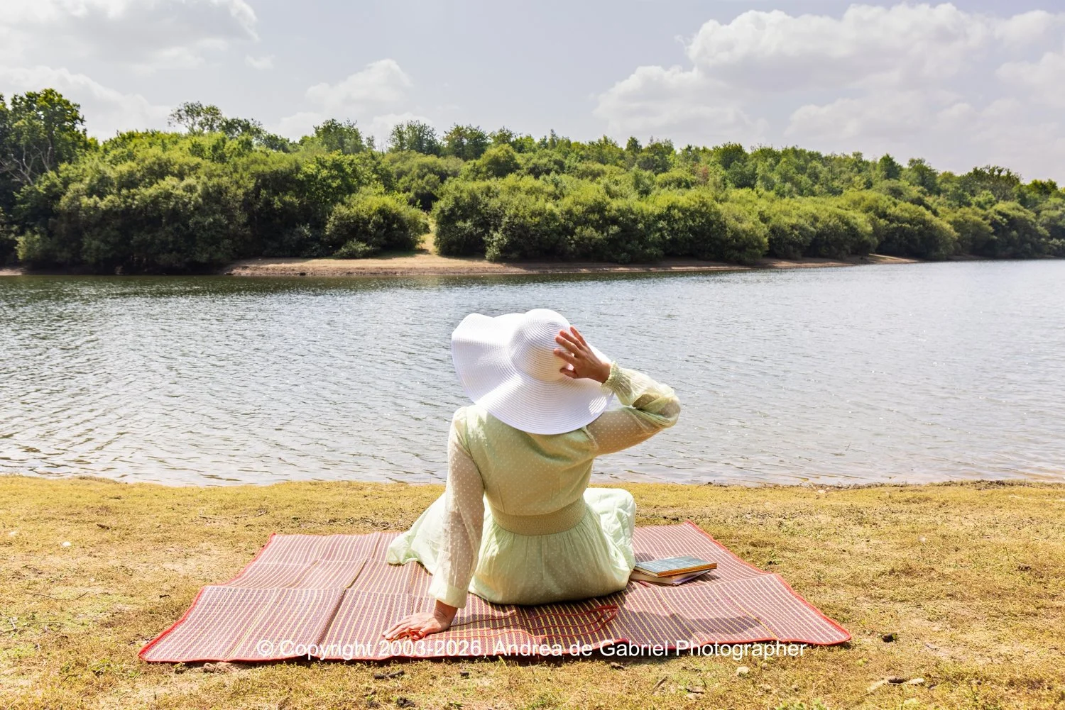 Woman sitting on a striped mat by a lake, wearing a large white sunhat and light-colored dress, looking at the water with trees in the background under a cloudy sky.