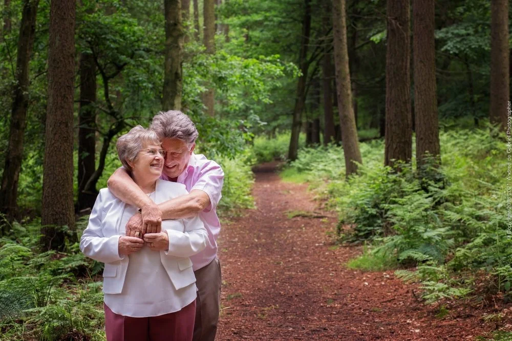 An elderly couple hugging and smiling in a forest with green trees and a dirt path.