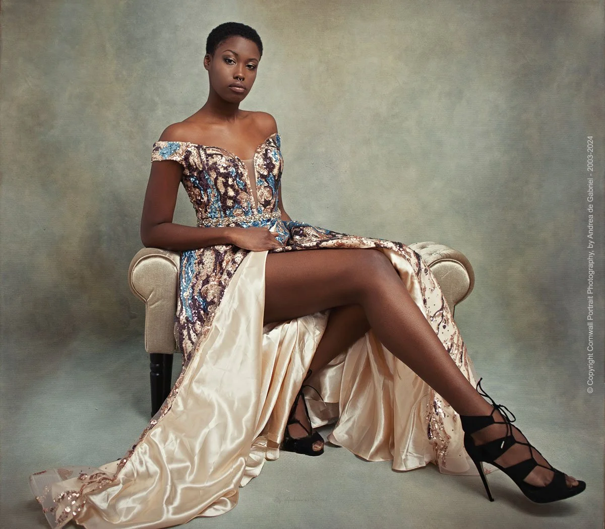 A woman sitting on a beige upholstered chair wearing an off-the-shoulder, sequined evening gown with a high slit, black high-heeled strappy shoes, and posing with her legs crossed against a neutral background.