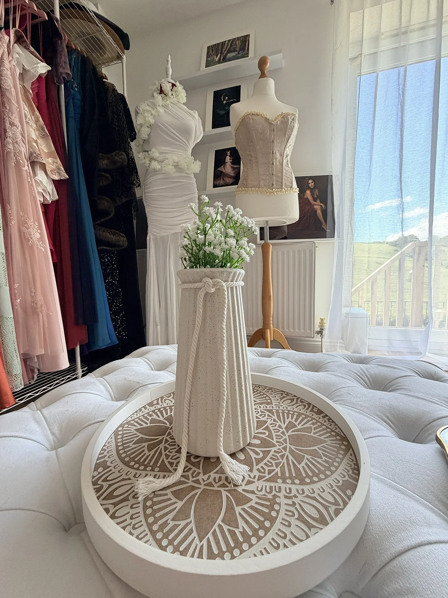 A white round table with a decorative top featuring a floral pattern, in front of a white tufted sofa. On the table, there is a white ceramic vase with a bouquet of white flowers. Behind the table, there are two dress forms displaying elegant dresses