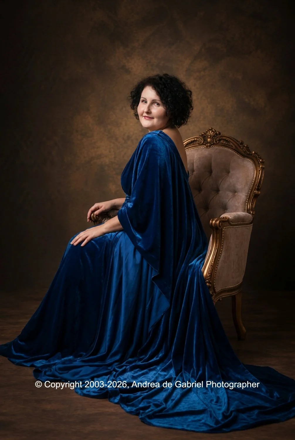 A woman with short, curly dark hair sitting on an ornate, vintage armchair. She is wearing a luxurious, deep blue velvet gown that drapes elegantly around her. The background is a warm, textured brown.