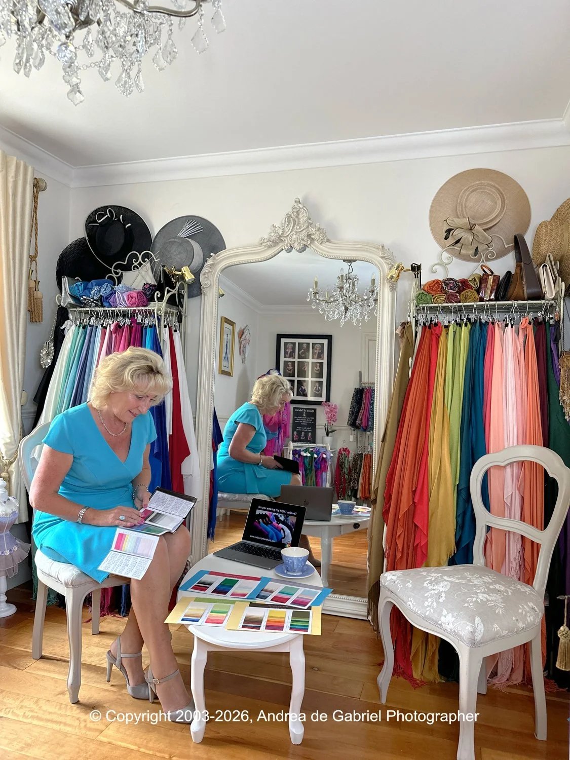 A woman in a blue dress reading a color fan book sitting on a white chair inside a fabric shop with colorful fabrics hanging on racks, a mirror, and a chandelier.