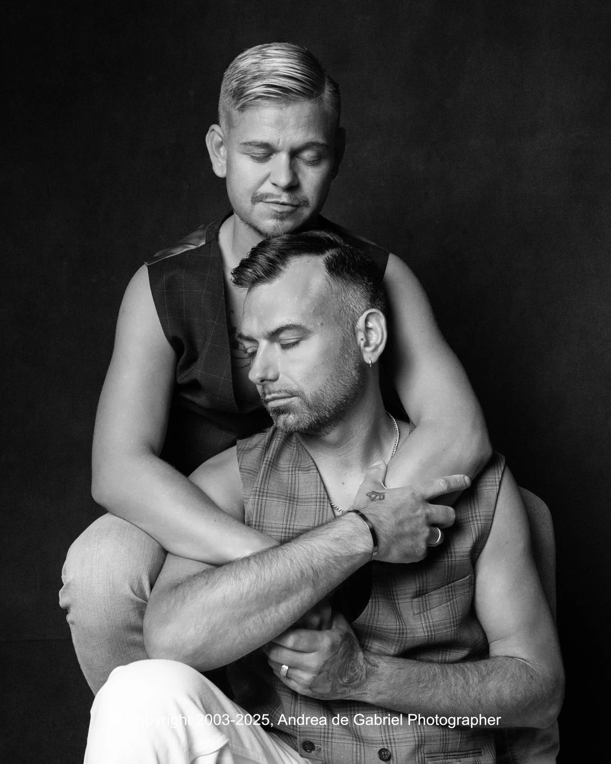 A black-and-white portrait of two men with closed eyes. One man is sitting and the other is standing behind him, embracing him with arms around his shoulders. Both men have short hairstyles, and the sitting man has tattoos on his arm.