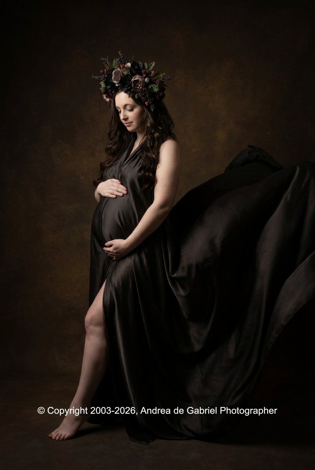 A pregnant woman with long dark hair in a dark satin dress, wearing a large floral crown, gently touches her belly with both hands, standing against a dark brown background.