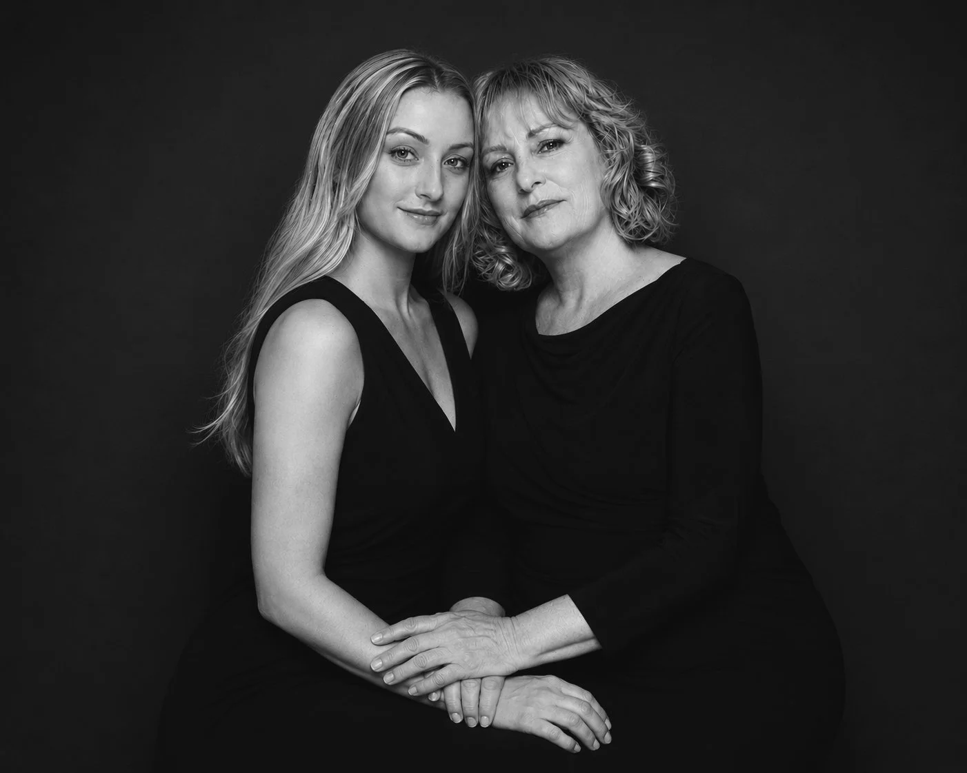 Black and white portrait of a young woman and an older woman sitting close together, both wearing black tops, with their hands gently clasped, against a dark background.
