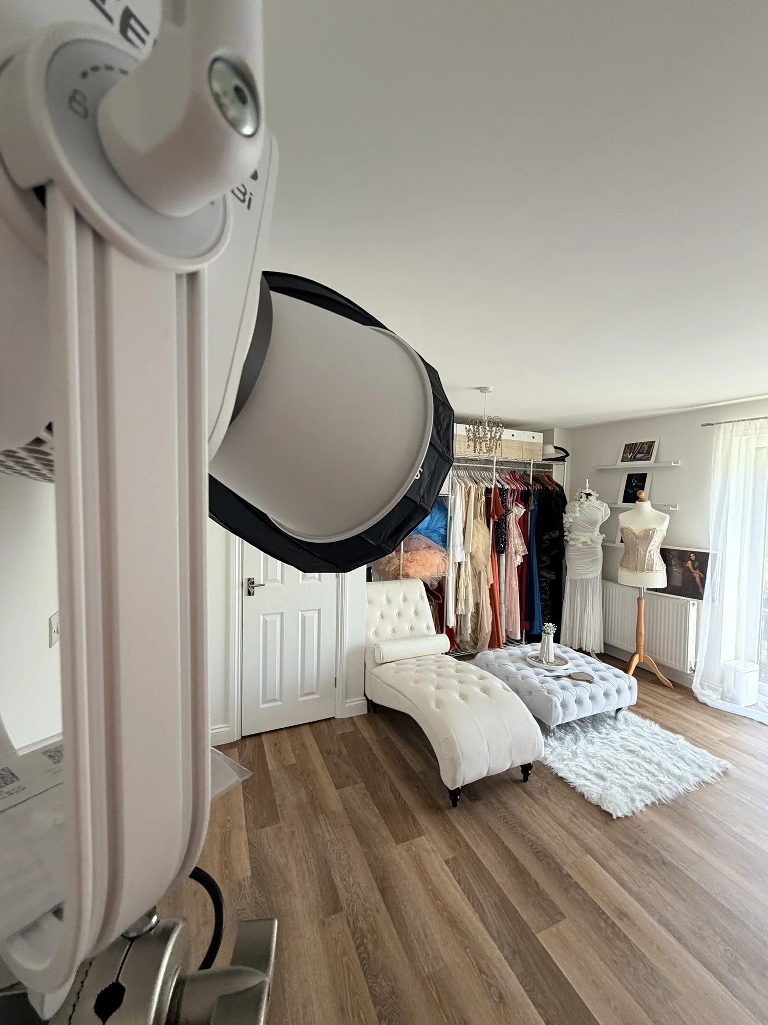 Photo of a room with clothing racks, a white tufted chaise lounge, a mannequin with a corset, and a fluffy white rug, taken from an angle showing part of a makeup mirror.