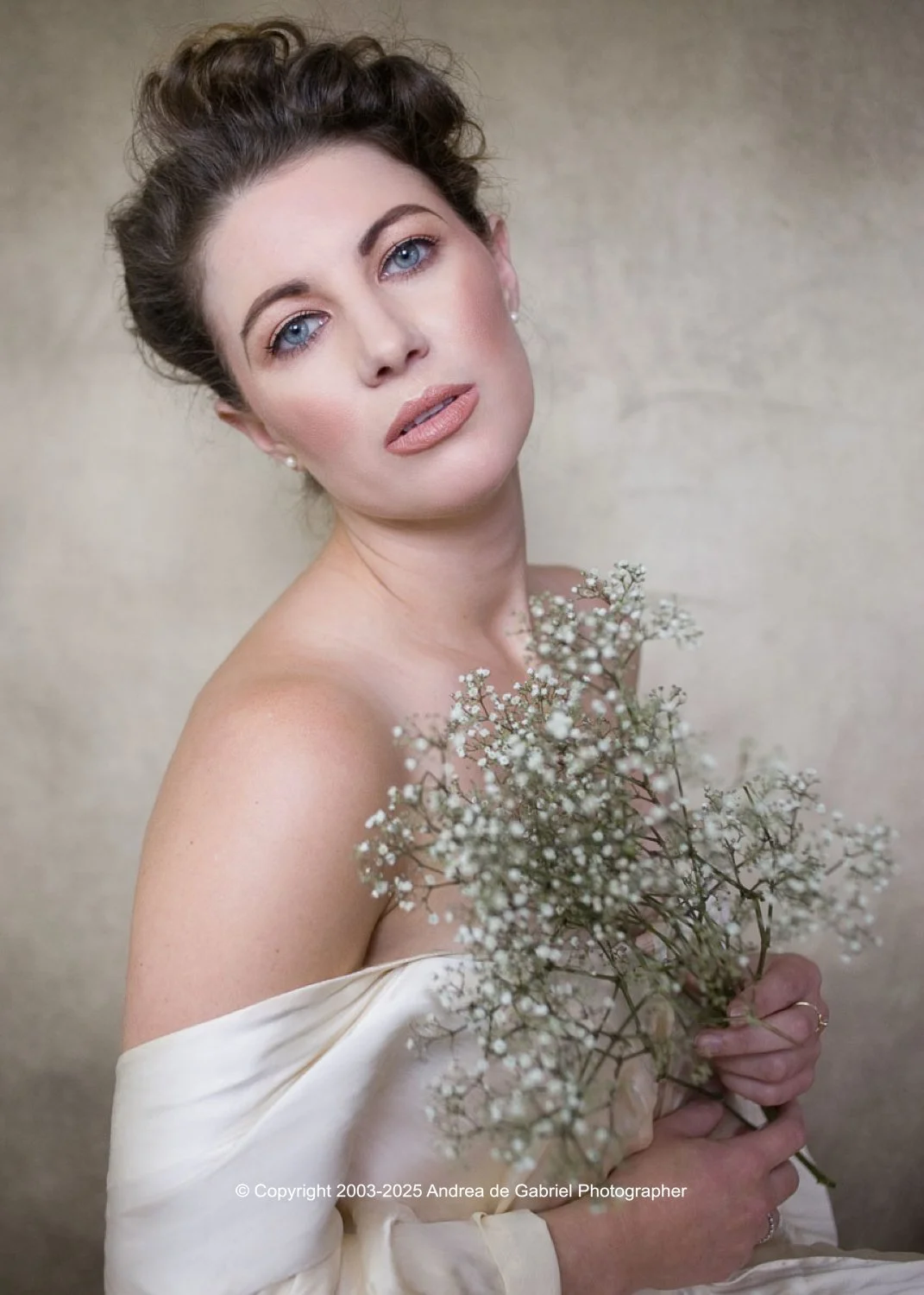 A woman with curly brown hair and blue eyes holding a bouquet of small white flowers, wearing an off-shoulder satin dress, and looking at the camera.