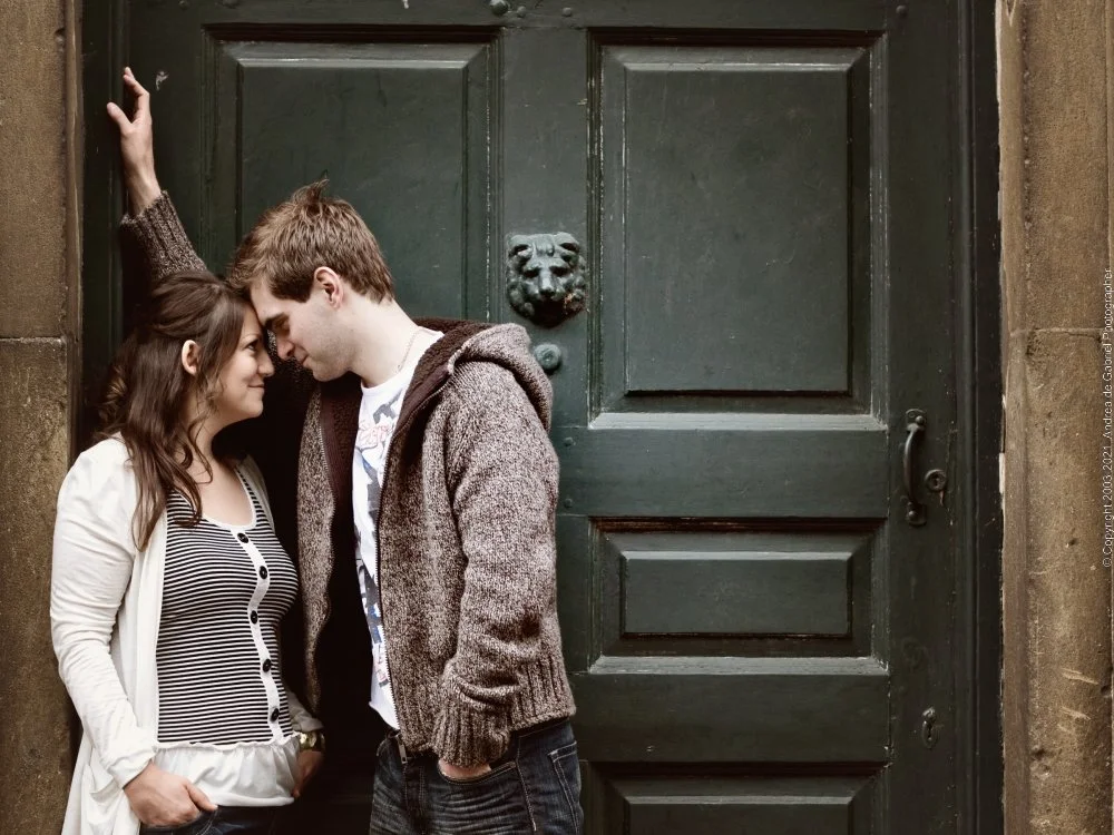 A young couple standing close together, touching foreheads and smiling, leaning against a green door with a lion's head door knocker.