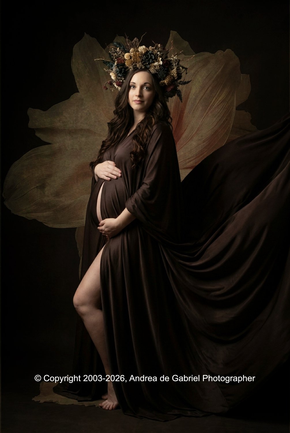 A pregnant woman wearing a brown satin dress, standing with one hand on her belly, and the other hand supporting her lower abdomen. She has long dark hair and wears a floral crown made of dried flowers. The background features large dried leaves and 