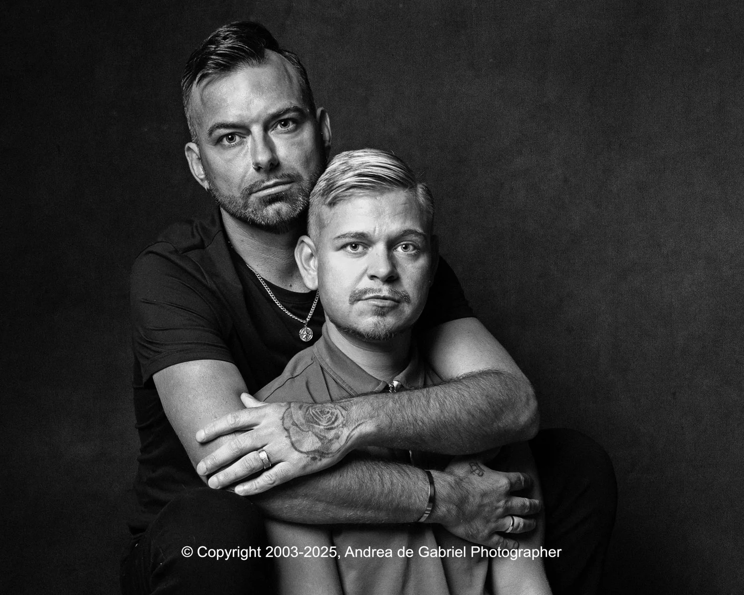 LGBT+ couple posing together in a black and white portrait against a dark background. One man is sitting and the other man is standing behind him, embracing him with his arms around his shoulders. Both have composed expressions.