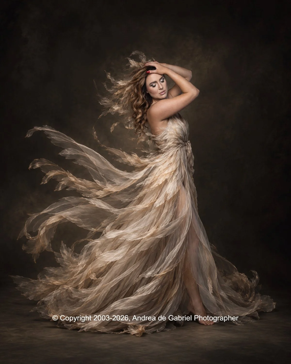 A woman in a flowing beige gown made of layered, feather-like fabric, with her eyes closed and arms raised, holding her head. The background is dark, and the dress appears to be moving as if the woman is dancing or in motion.