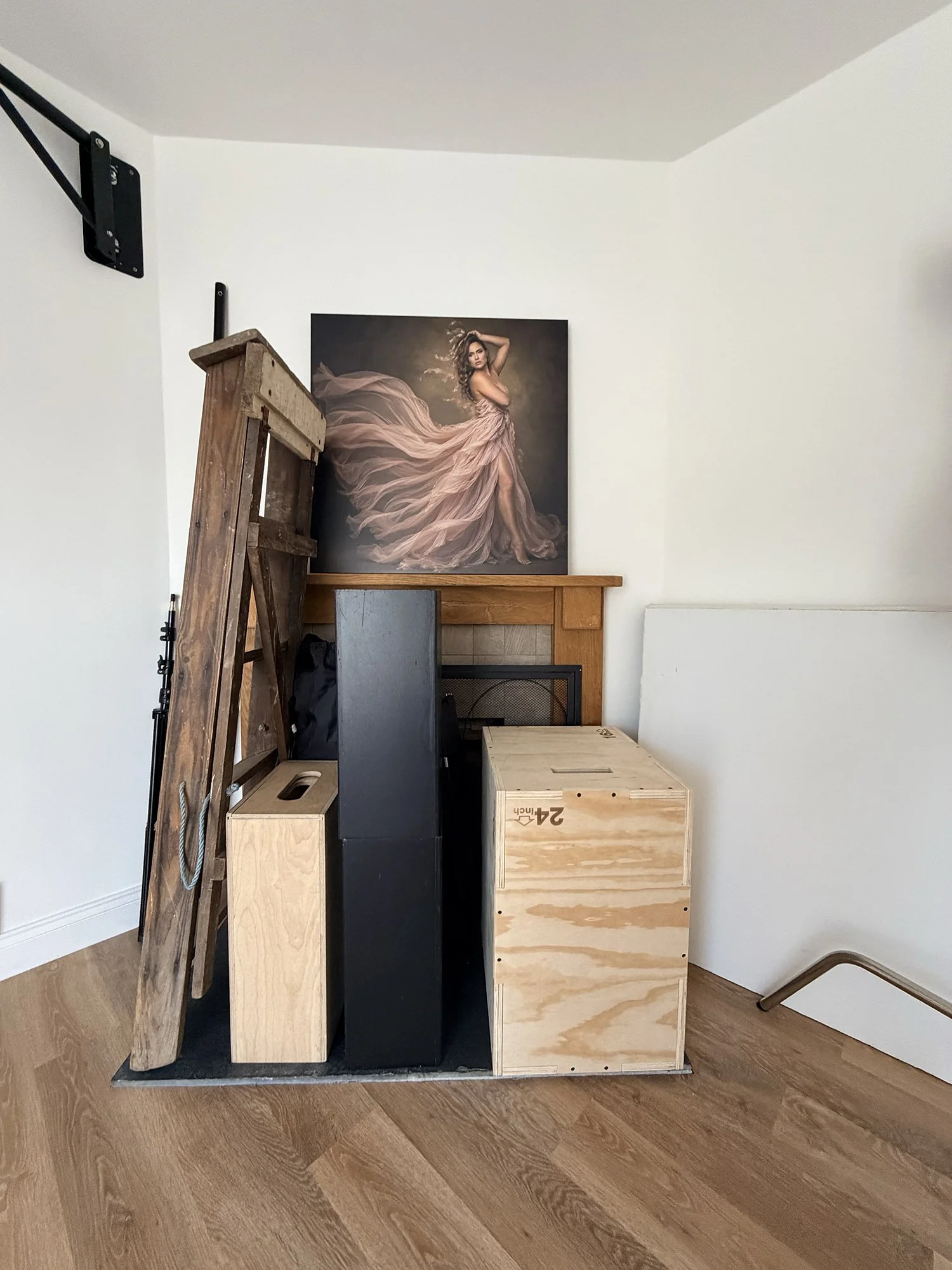 Various pieces of furniture, including a wooden kitchen island, a black rectangular block, a wooden box, and a large black and white photo of a woman in a flowing dress, all placed on a hardwood floor in a room with white walls.