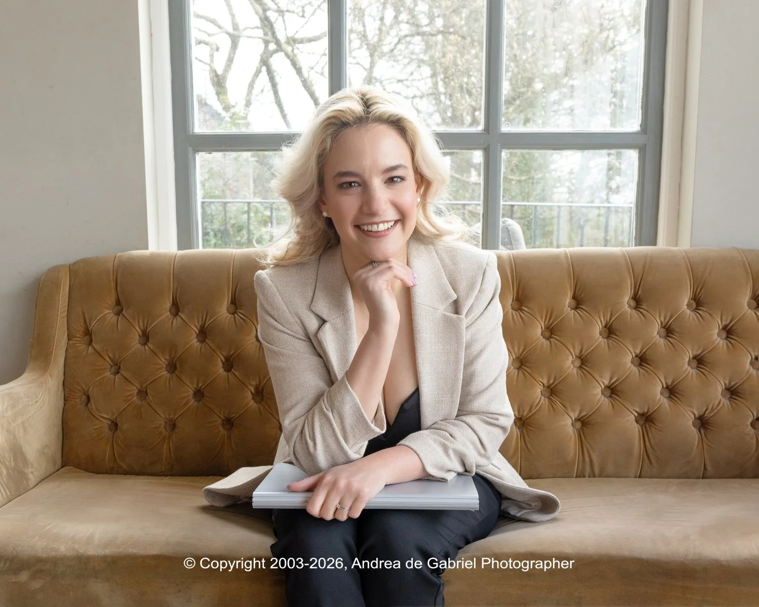 A smiling woman with blonde hair wearing a beige blazer, holding a notebook, seated on a tan tufted sofa in front of a large window with trees outside.