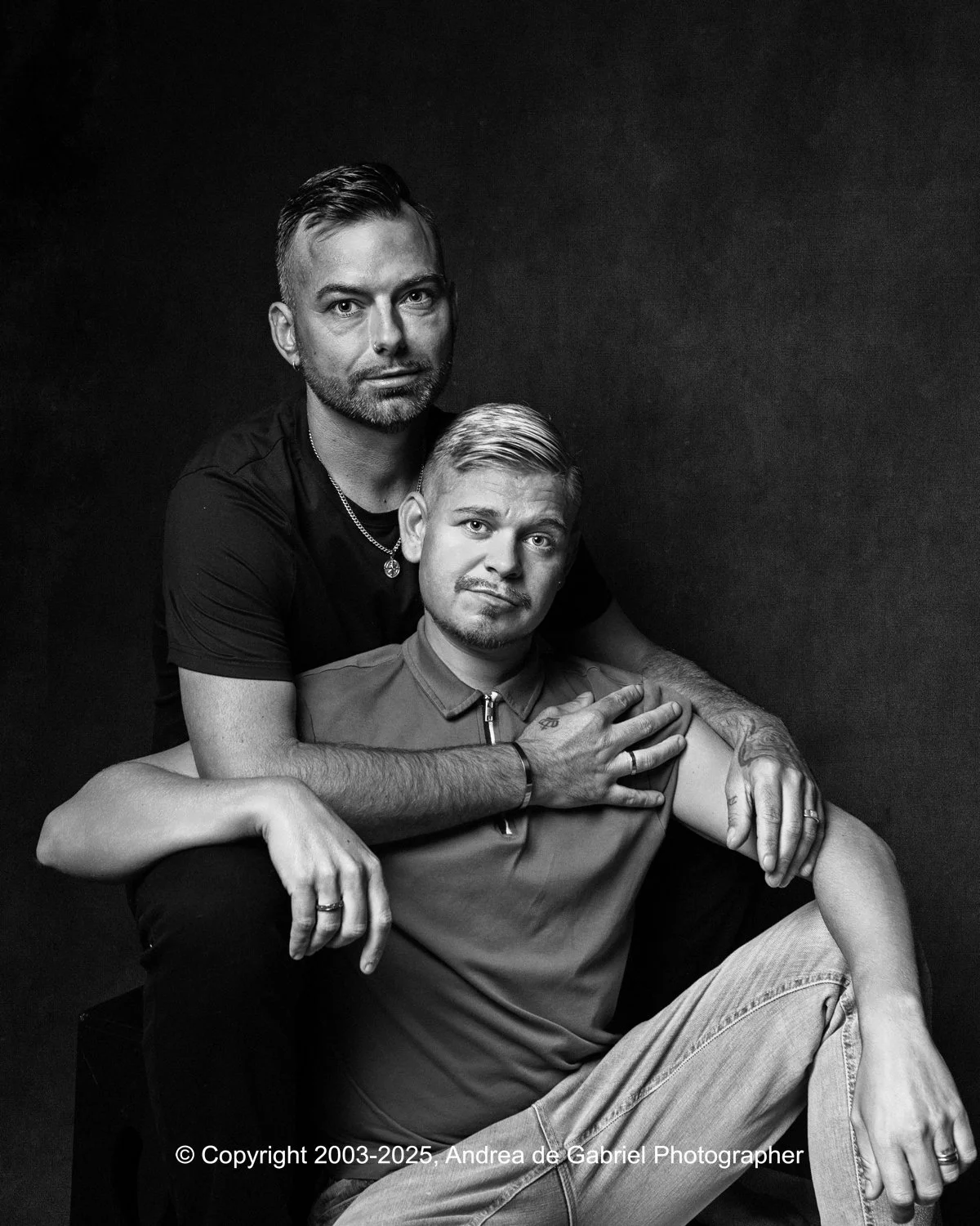 Gay male couple posing together in a black and white photo, with one sitting and the other standing behind him, both looking at the camera.