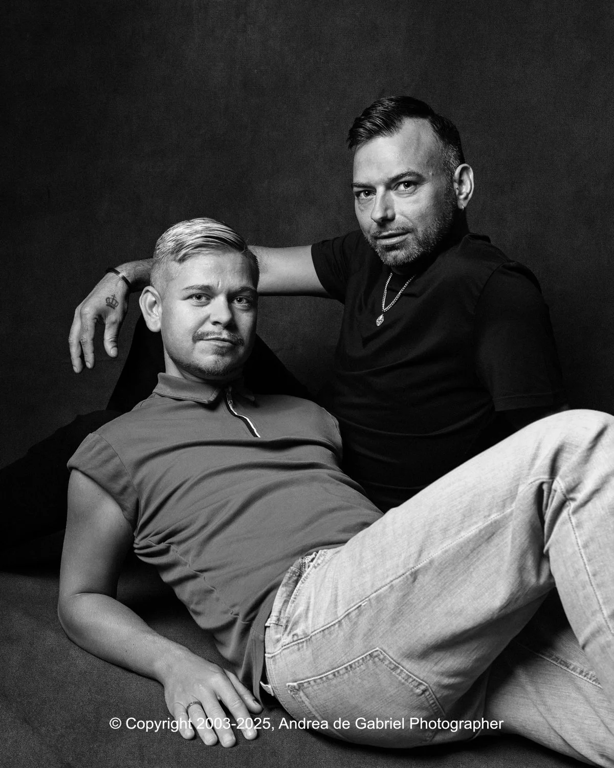 Two men posing together in a black and white photo, one sitting and one leaning on him, against a dark background. LGBTQ+ Couples' Portrait Photography in Cornwall, Devon and South West.