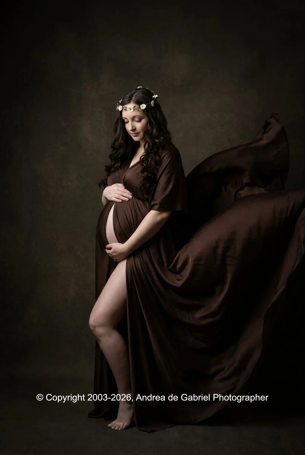 Pregnant woman in a brown dress with floral headband, gently holding her baby bump, against a dark background.