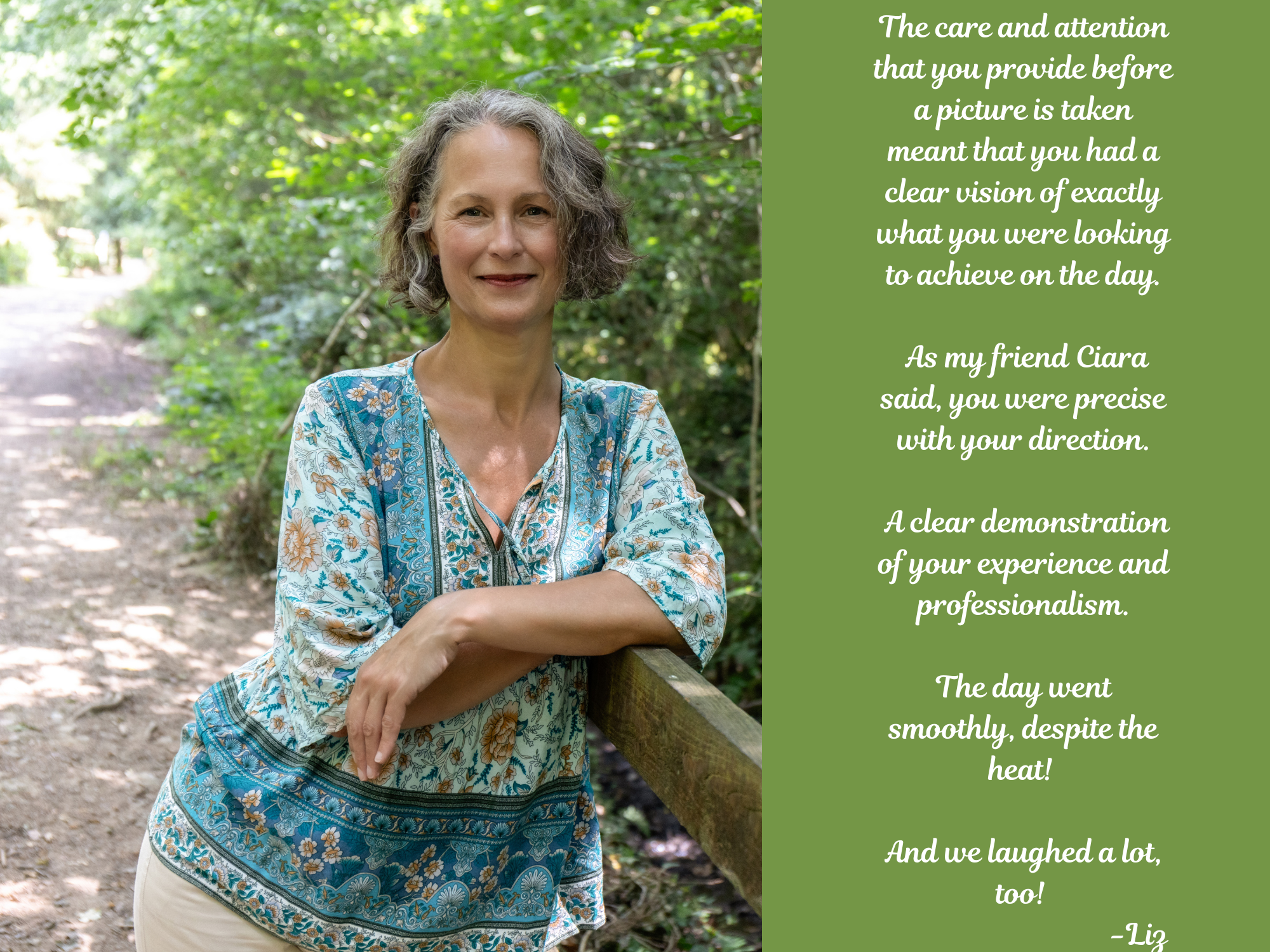 A woman with curly gray hair standing outdoors on a wooded path, leaning against a wooden railing, wearing a blue and white patterned blouse, smiling with arms crossed, surrounded by green foliage.