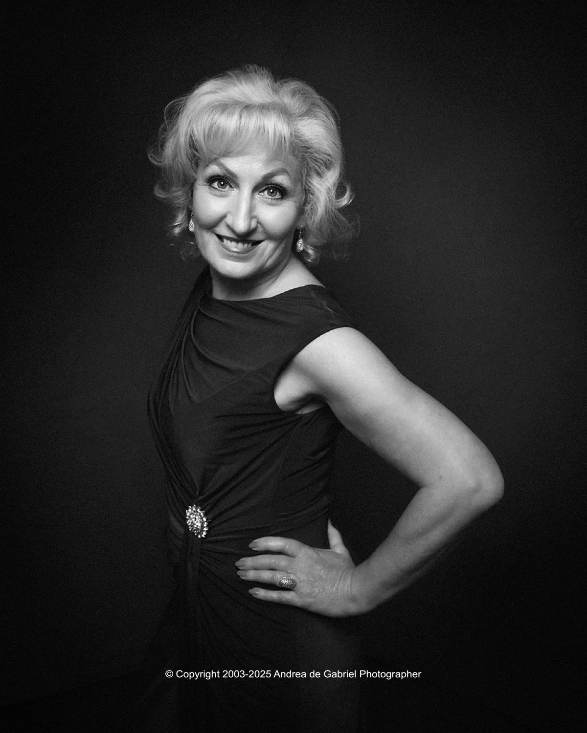 A black and white portrait of an older woman with styled hair, wearing a sleeveless dress with a brooch, earrings, and a ring, smiling and posing with one hand on her hip, against a plain dark background.