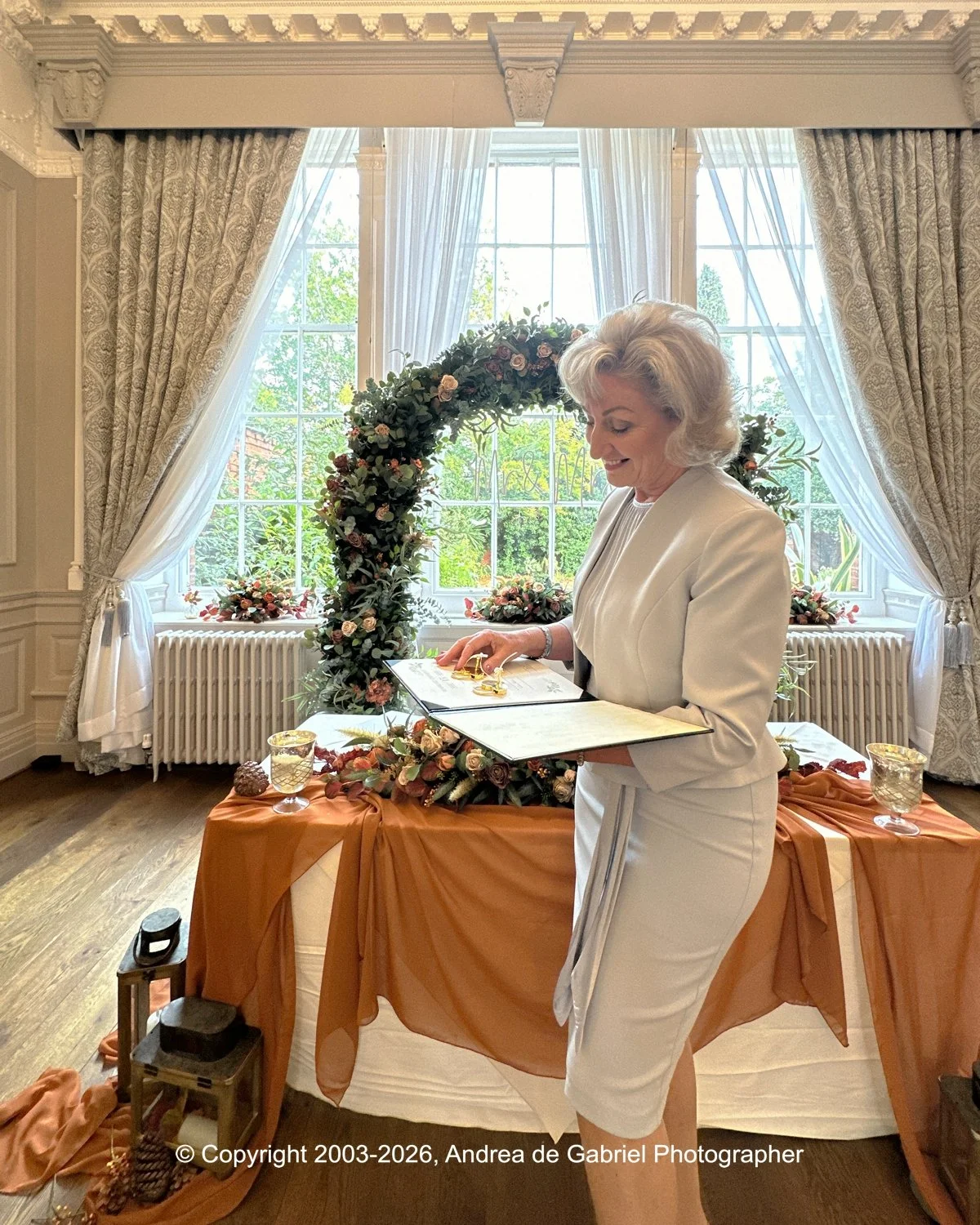 A woman in a white suit signing a document at a decorated table with floral arrangements, in a room with large windows, ornate curtains, and a floral arch backdrop.