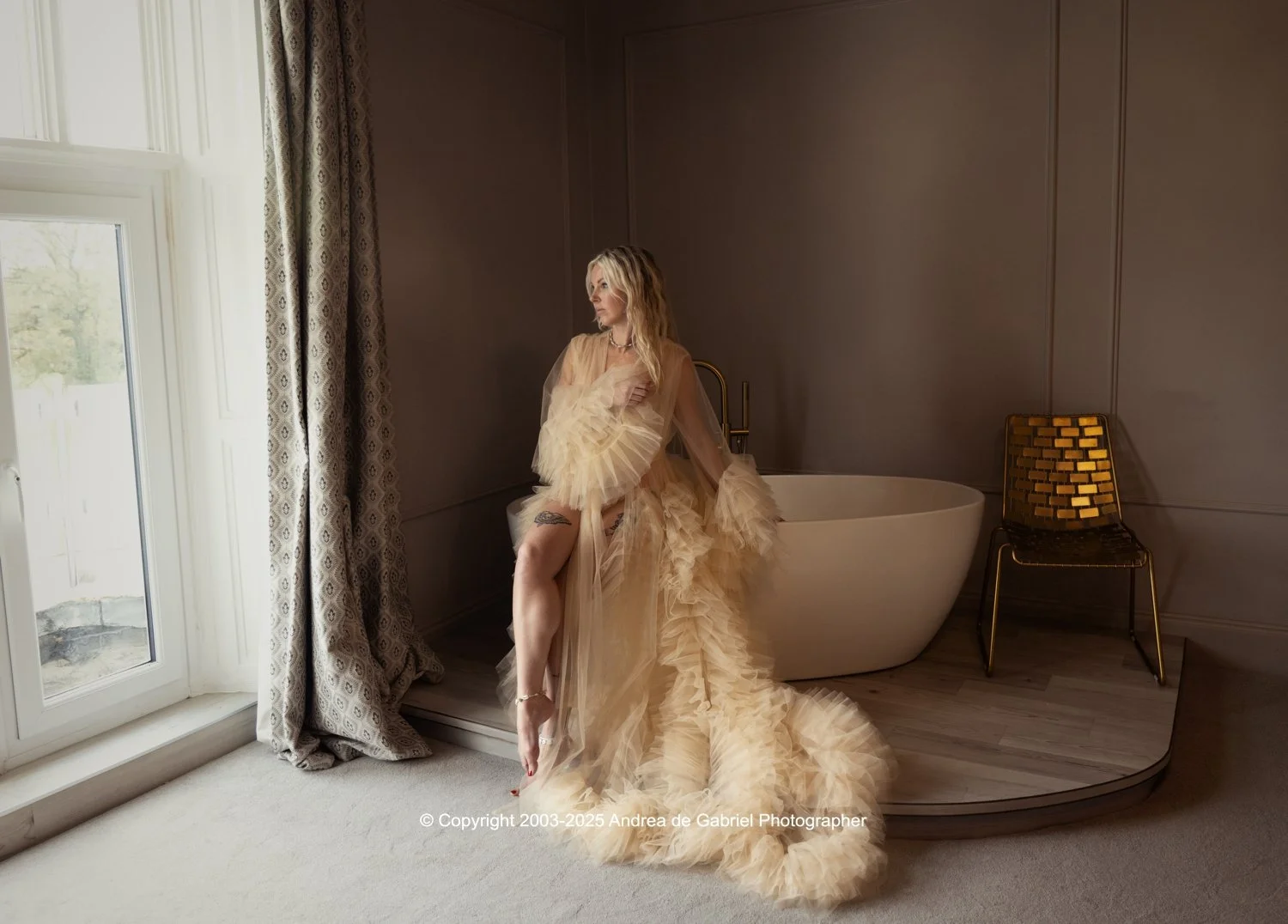 A woman in an elegant beige tulle gown with ruffles, standing next to a bathtub near a window with patterned curtains in a bathroom or spa setting.