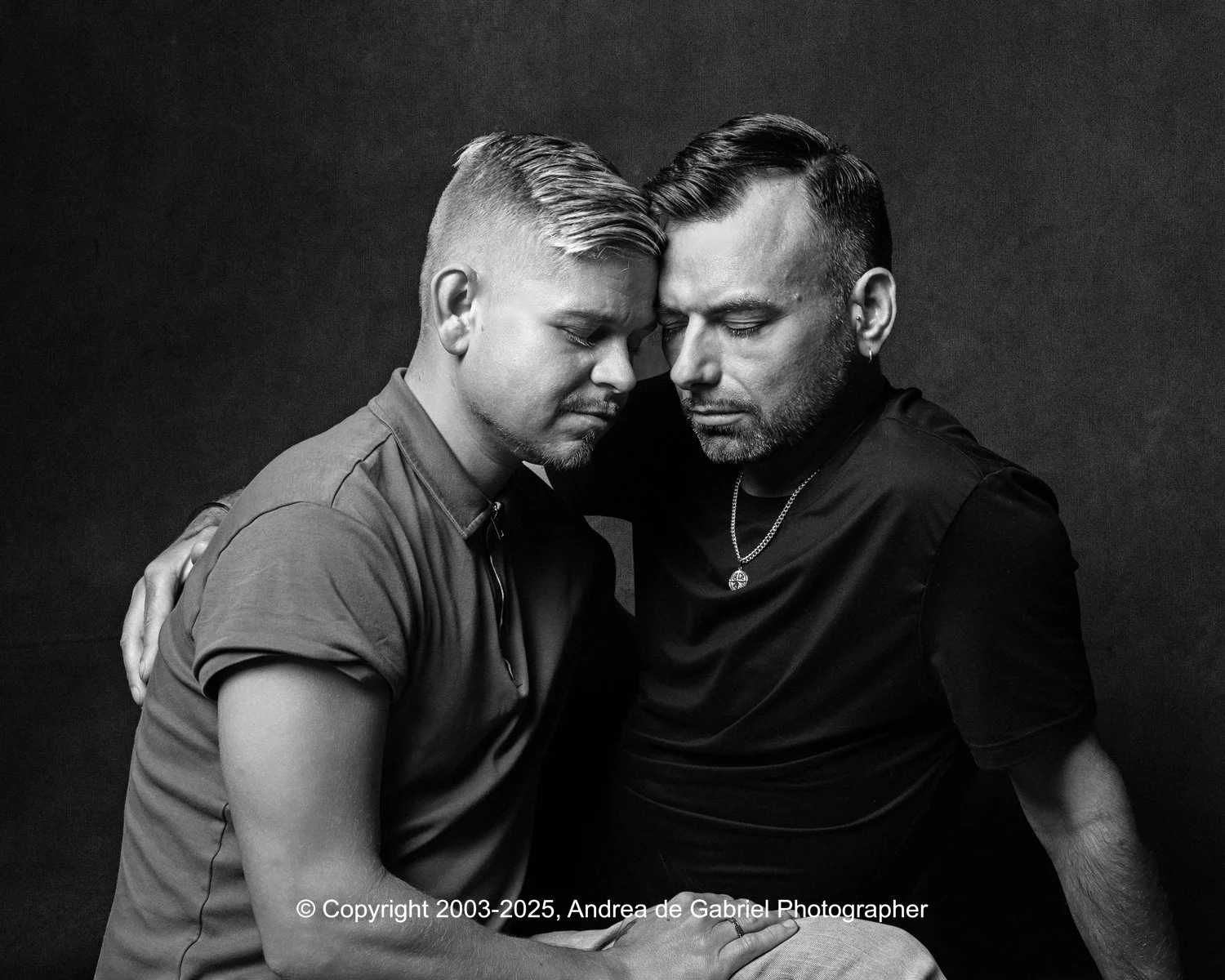 Same sex couple sitting closely with foreheads and noses touching, eyes closed, conveying intimacy and comfort in a black and white photo.