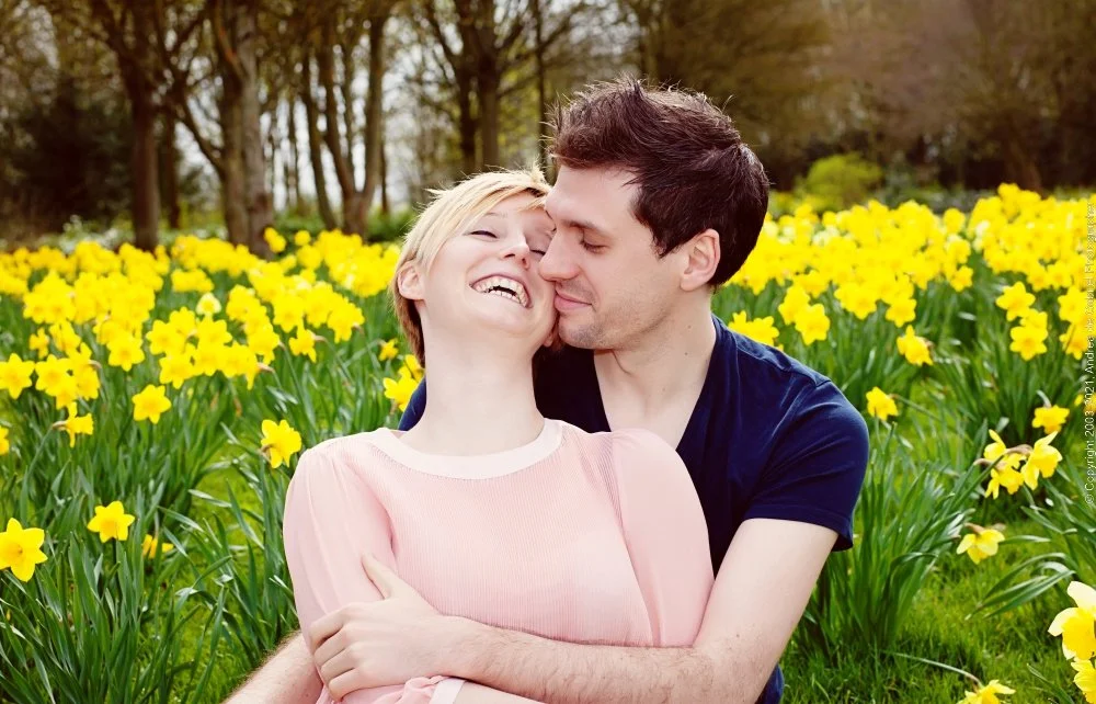 A couple hugging and smiling in a field of yellow flowers with trees in the background.