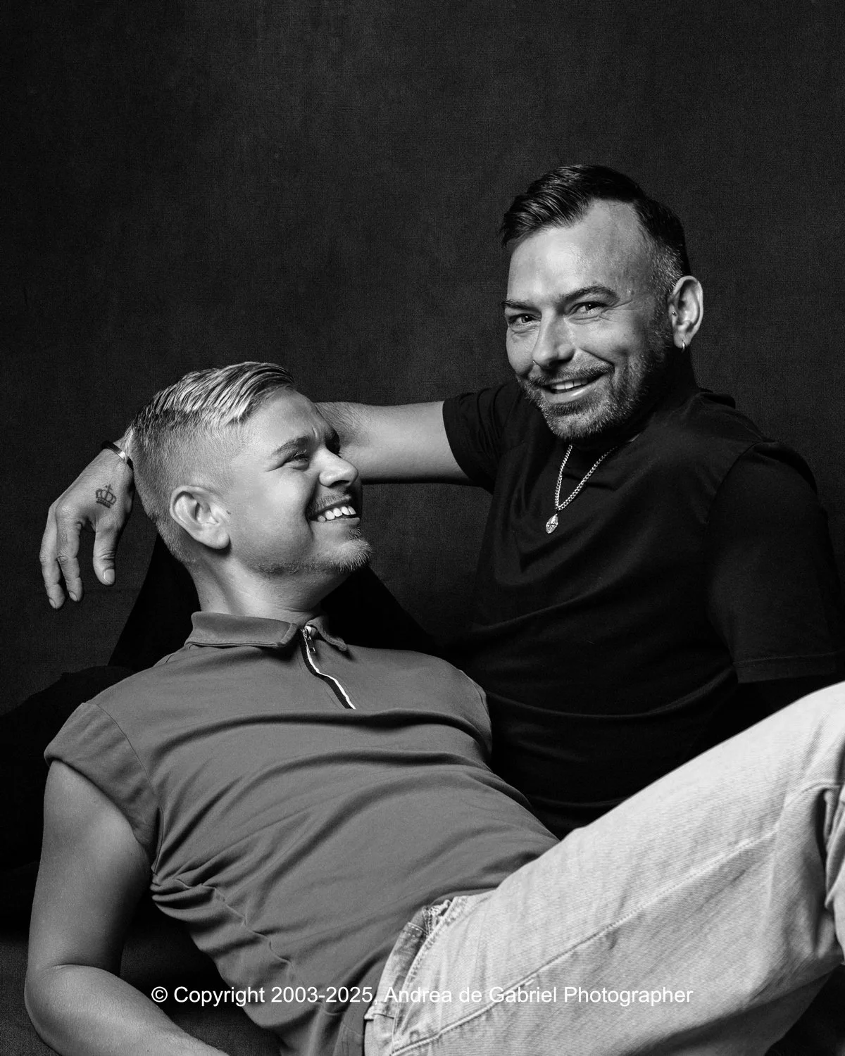 Same sex couple smiling and laughing together, one lying down and the other sitting with his arm around the other, black and white photo.