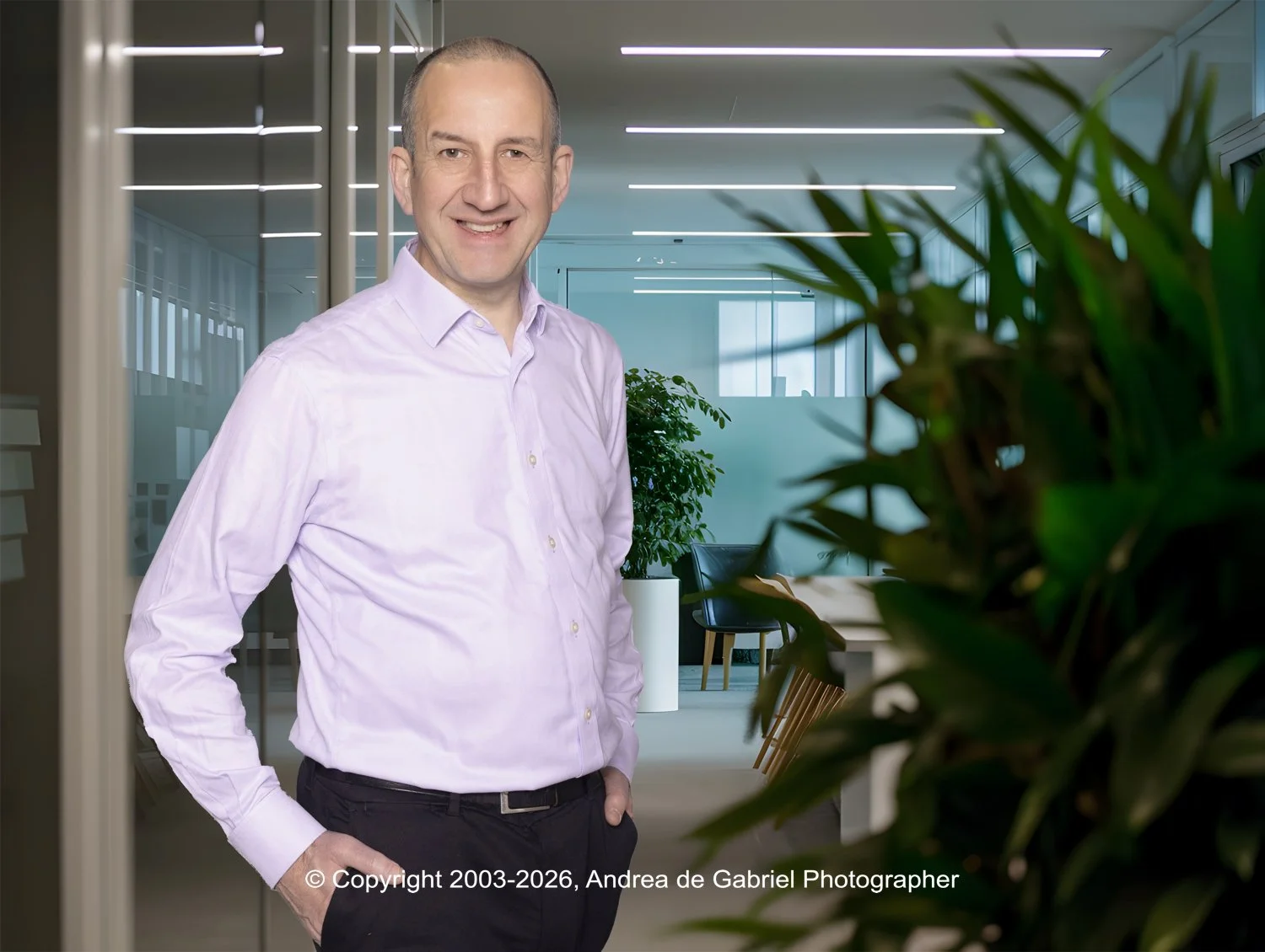 A man in a light pink dress shirt and black pants standing in a modern office space with glass walls, large windows, and indoor plants.