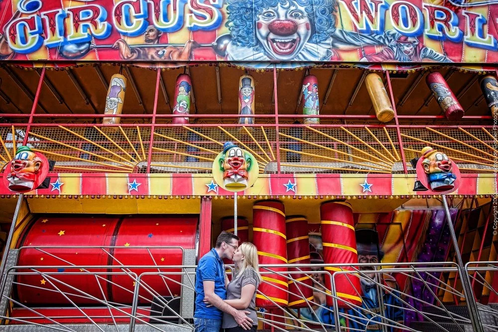 A couple kissing in front of a colorful carnival ride called Circus World. The ride features clown faces, striped decorations, and a large red barrel-shaped structure.