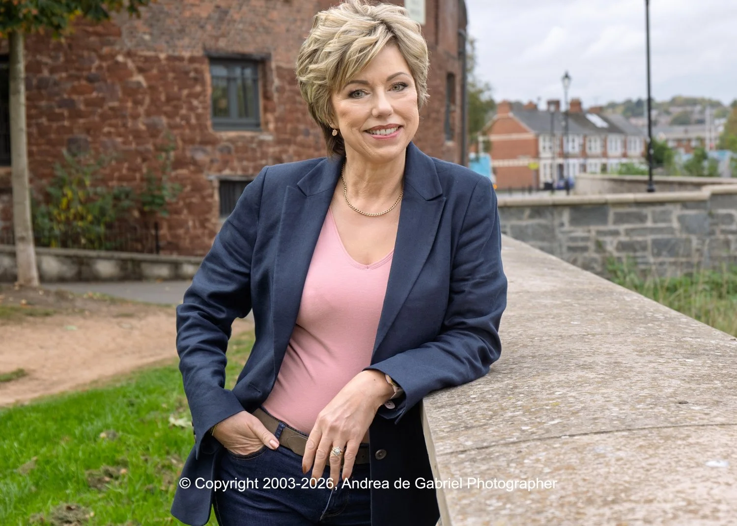 A smiling woman with short blonde hair wearing a navy blazer, pink top, and gold jewelry outdoors on a cloudy day, leaning on a stone ledge with a red brick building and distant houses in the background.