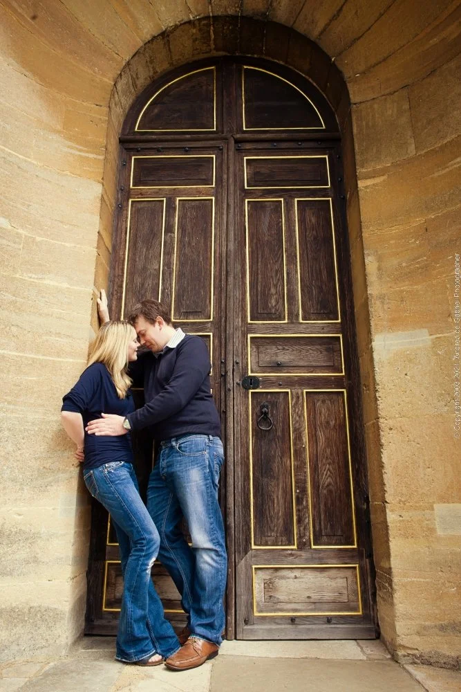 A couple standing close together in front of a large wooden door, with the woman leaning her head against the man's forehead, both appearing intimate.