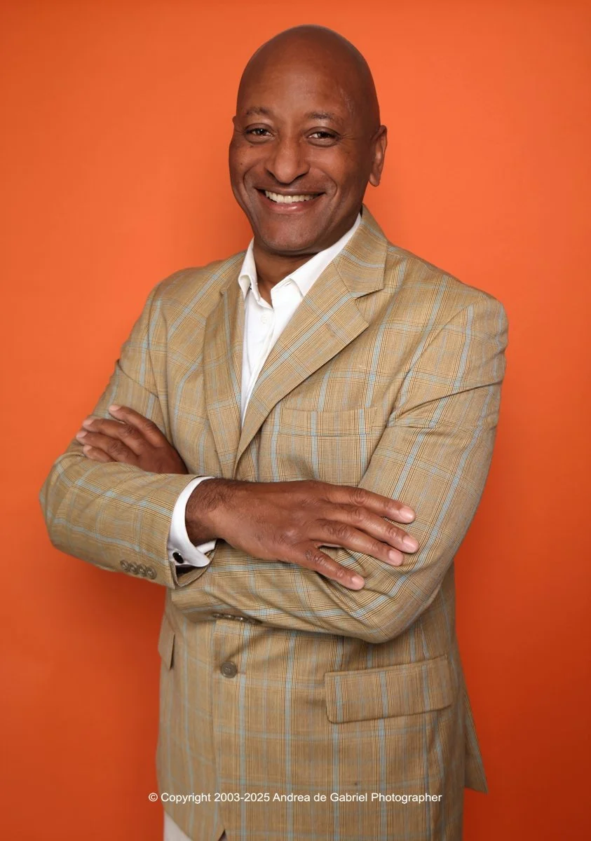 A smiling man with a bald head, wearing a beige plaid suit and white shirt, standing with arms crossed against an orange background.
