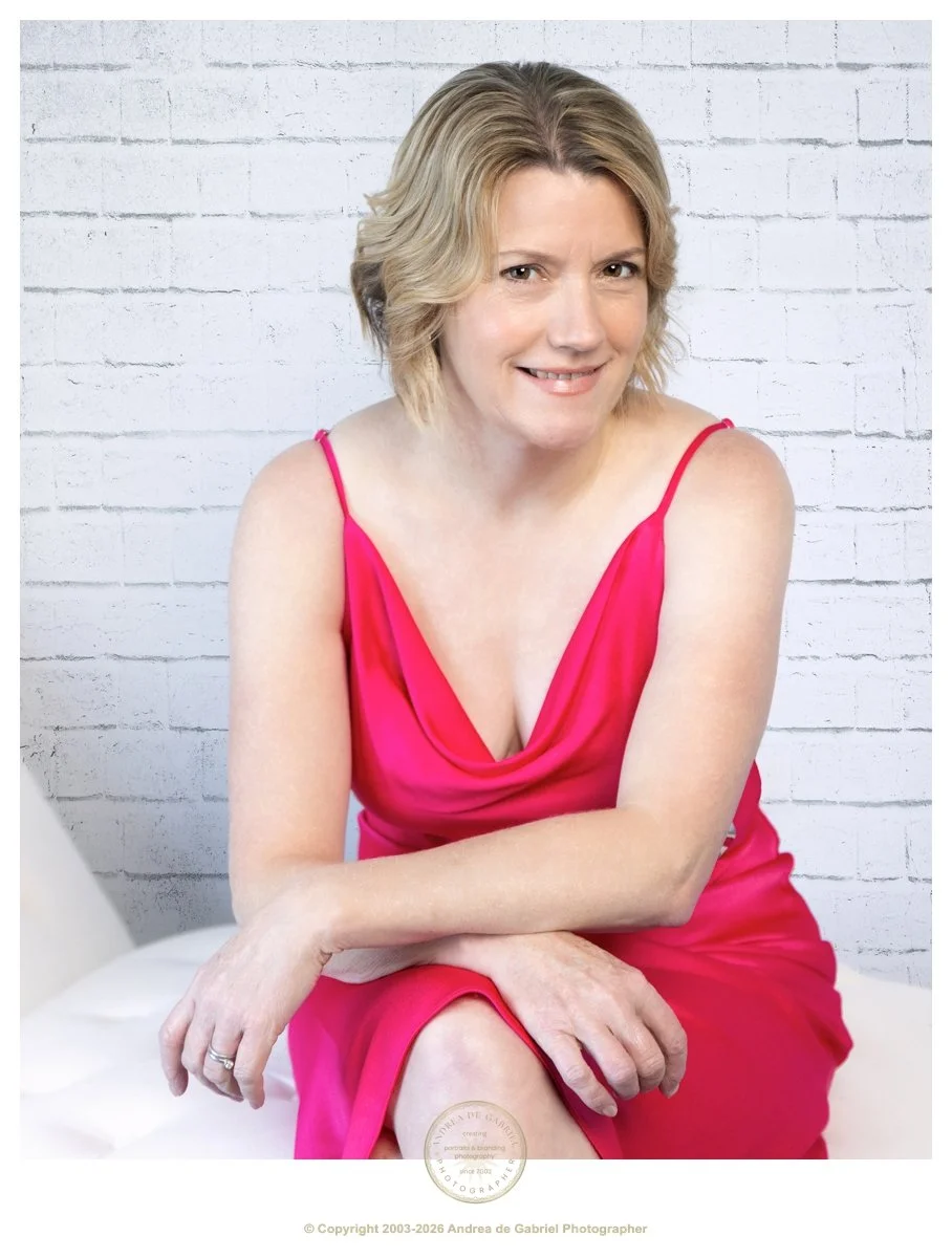 woman with short blonde hair, sits with crossed arms on her legs, facing the camera. she is wearing a pink dress, and smiling.