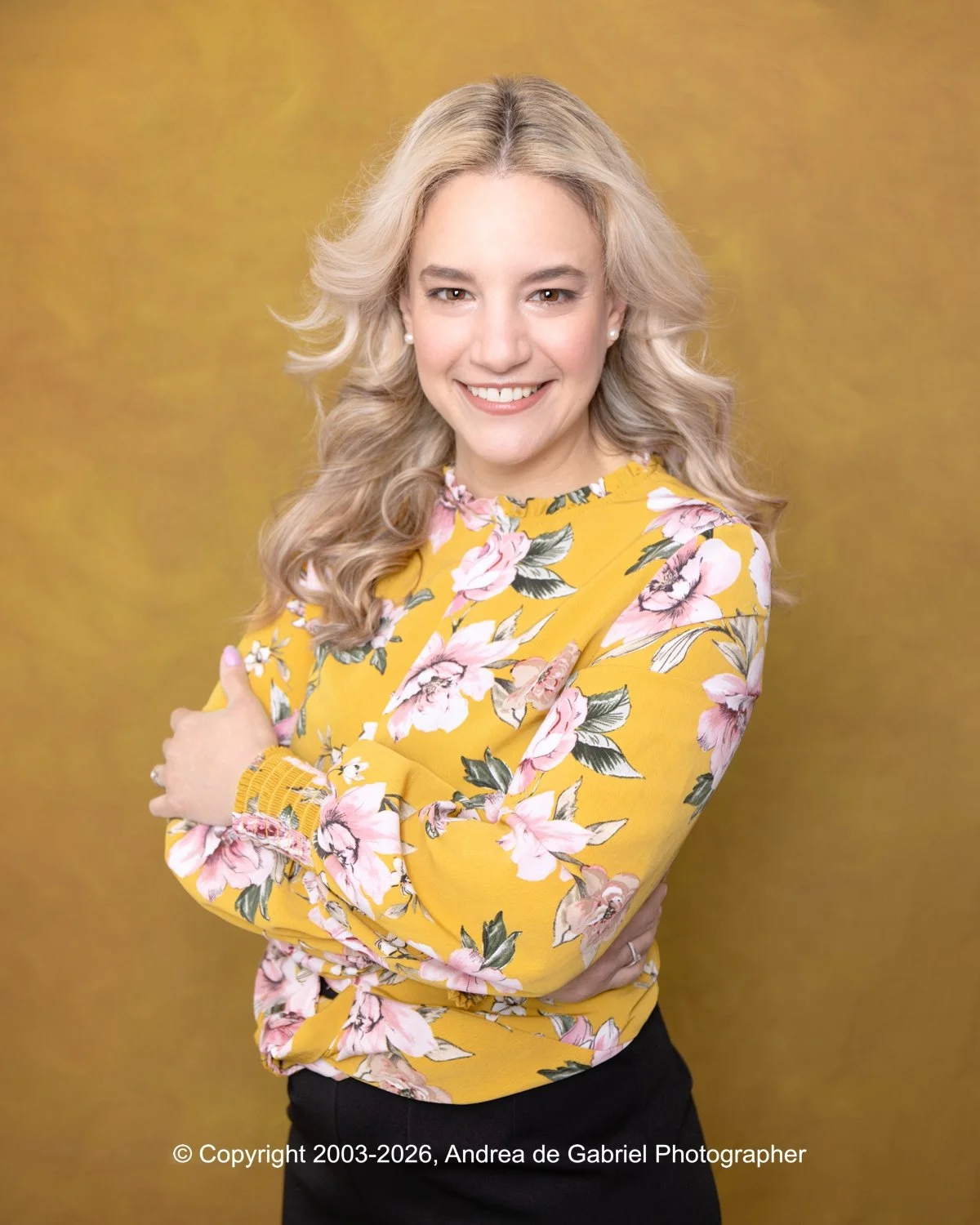 A woman with long wavy blonde hair smiling, wearing a yellow floral blouse and black pants, standing against a yellow background.