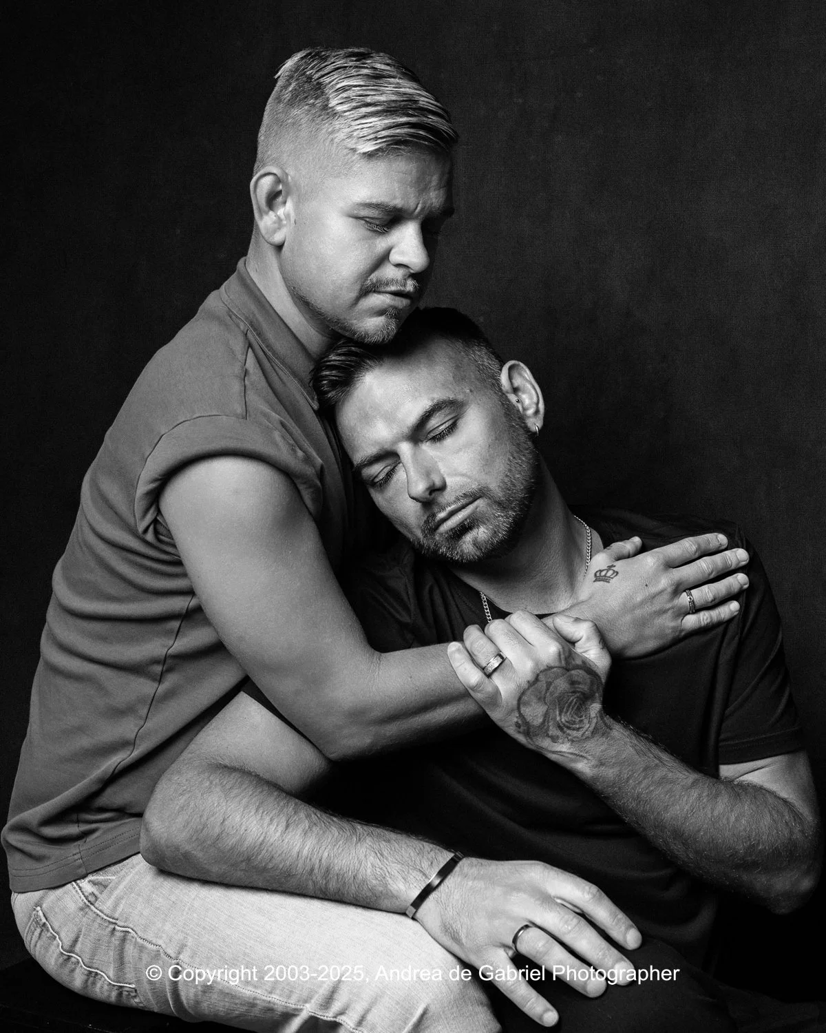Same sex couple married couple embracing in a black and white photo, one sitting with eyes closed and the other leaning over him, both with affectionate expressions.
