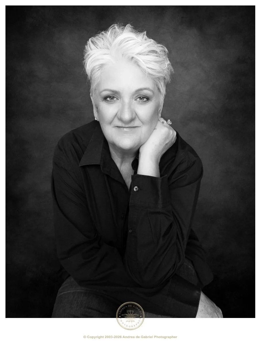 black and white image of mature woman, sitting and looking at the camera. she has a serene smile, and has white hair. she is wearing a black shirt and jeans.