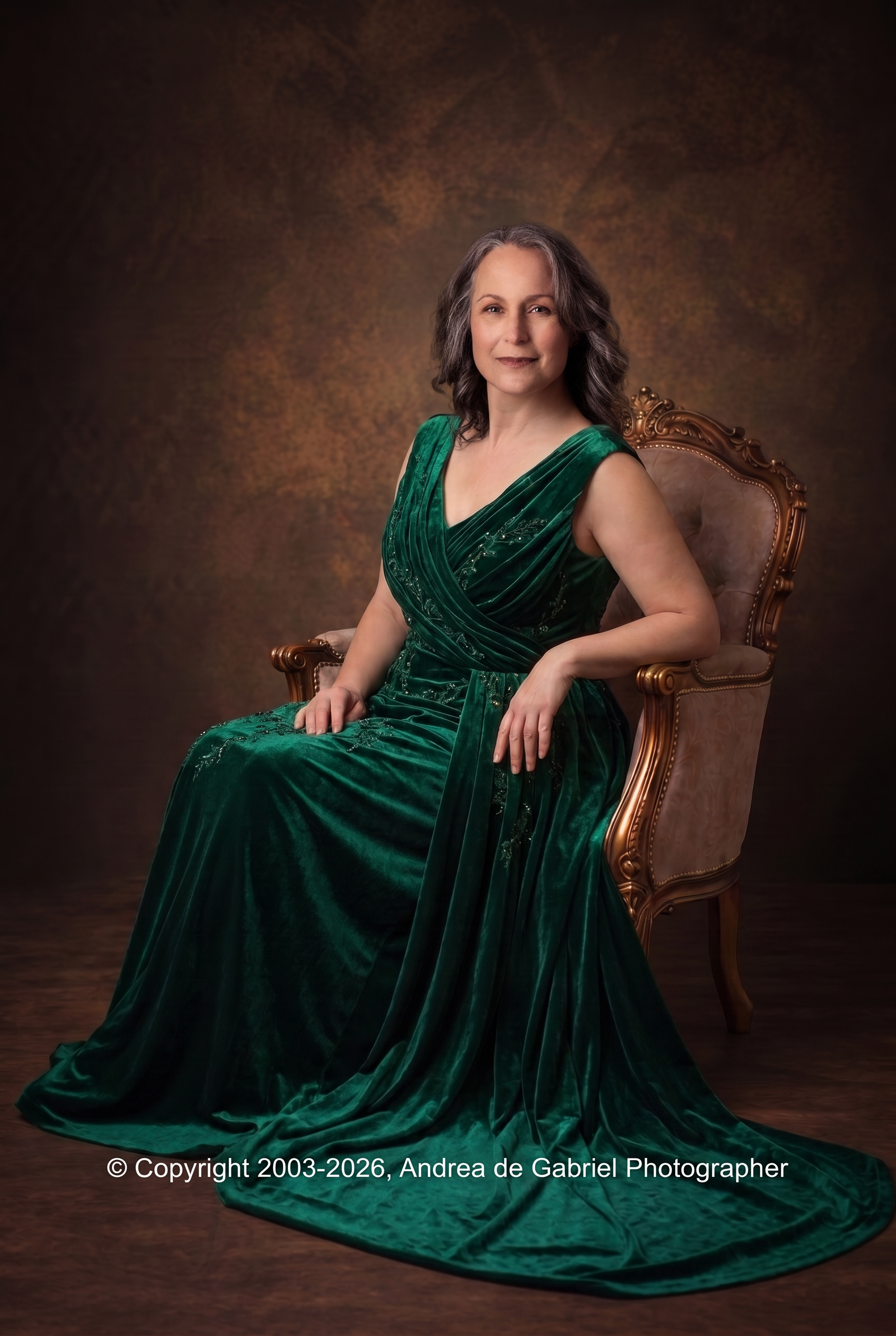 A middle-aged woman with gray curly hair and fair skin, sitting on an ornate wooden chair with gold accents, wearing a long, elegant forest green velvet gown, against a dark mottled background.