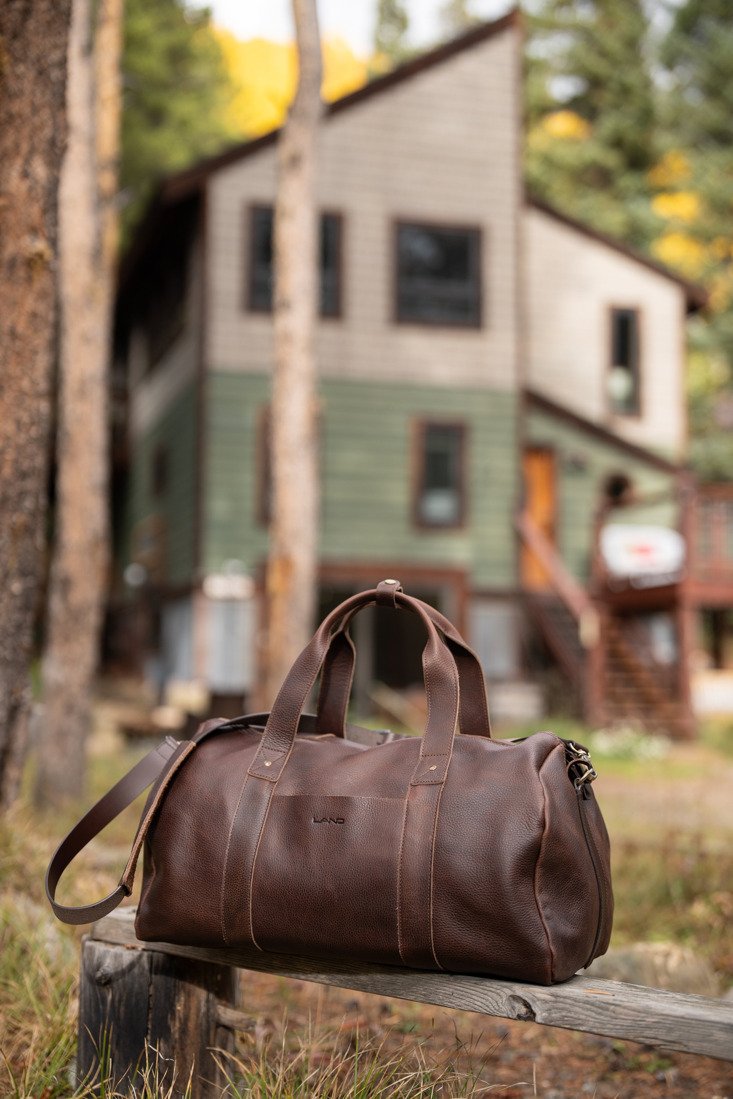 A brown leather duffel bag resting on a small wooden bench outdoors, with a blurred background of a two-story house with green and beige siding and trees.