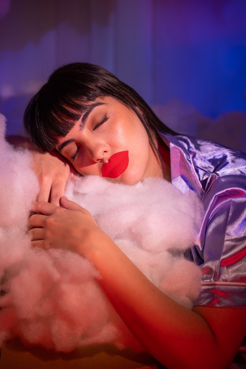 Woman sleeping on fluffy white pillow or blanket with lips-shaped red lipstick mark on her lips, wearing a shiny purple satin robe, in a colorful, soft-lit room