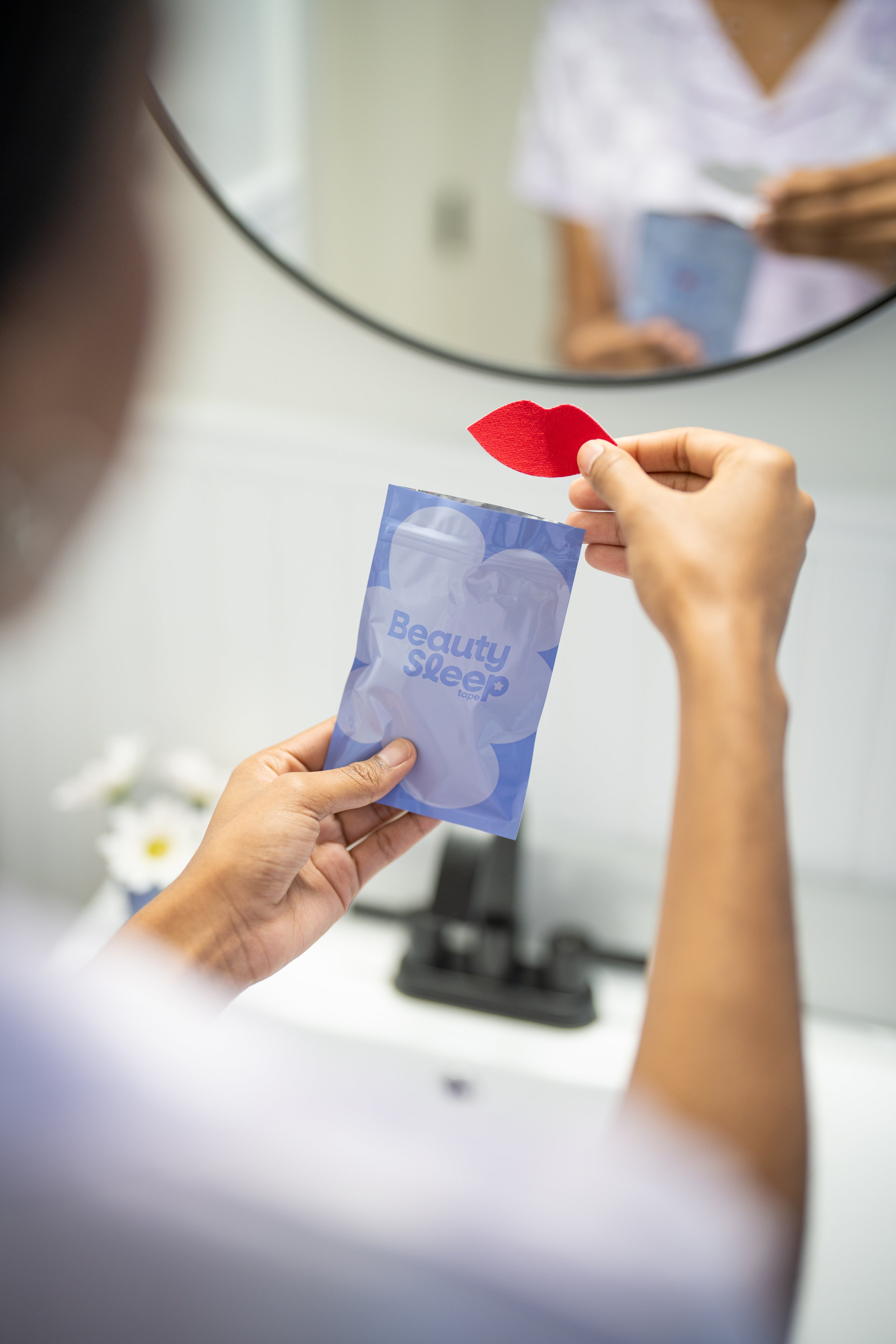Person holding a packet labeled 'Beauty Sleep' and a red sleep mask in front of a mirror.