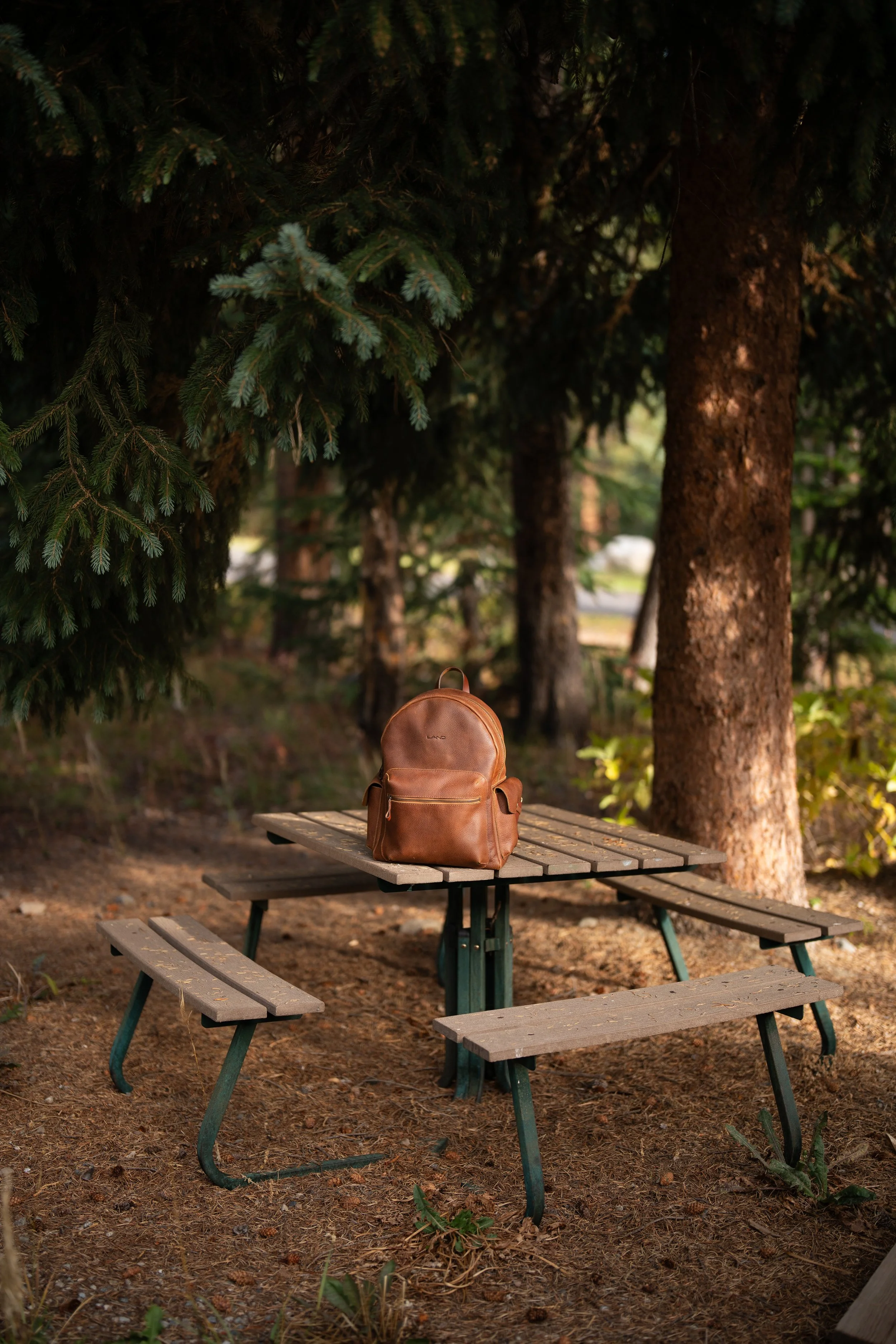 A brown leather backpack placed on a wooden picnic table outdoors, surrounded by trees and natural ground.