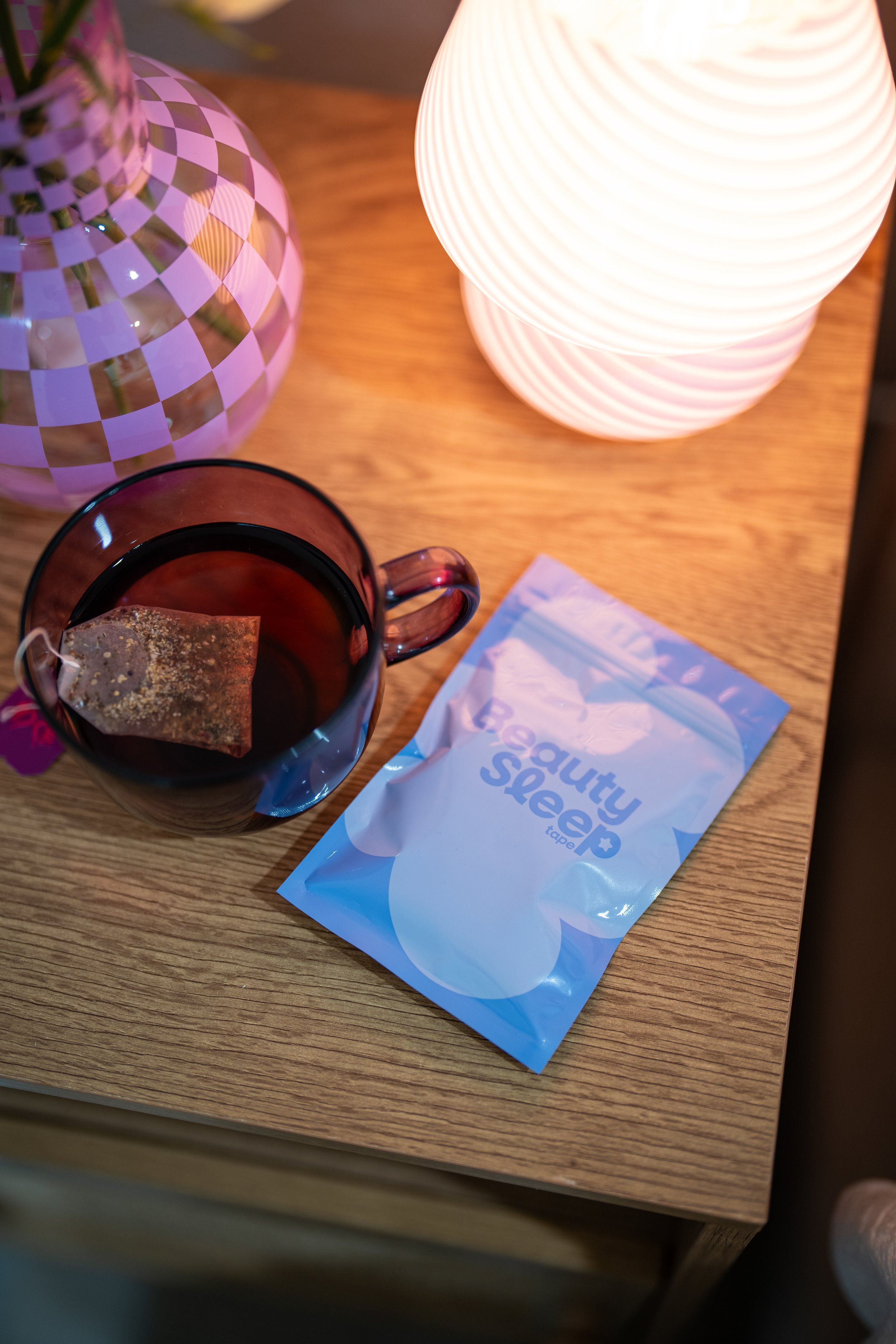 A wooden table with a lit white lamp, a checkered purple and clear glass vase, a cup of tea with a tea bag, and a package of beauty skin tape.