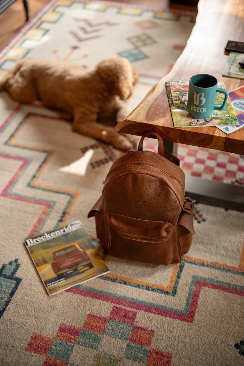 A brown leather backpack on a patterned area rug next to a wooden table, with a magazine on the floor showing a vintage truck, and a dog lying on the rug nearby.