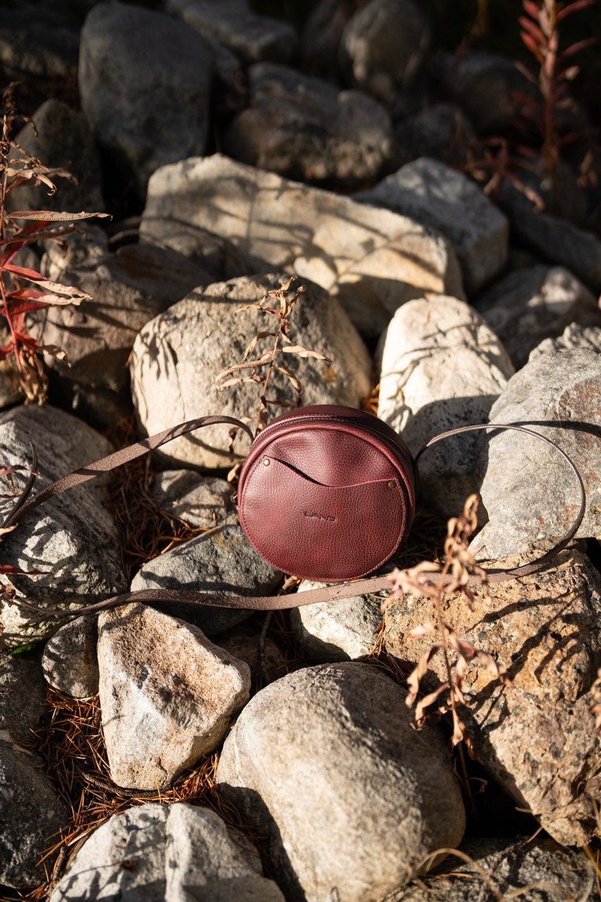 A small, round, burgundy leather crossbody bag resting on rocks and dried plants outdoors.