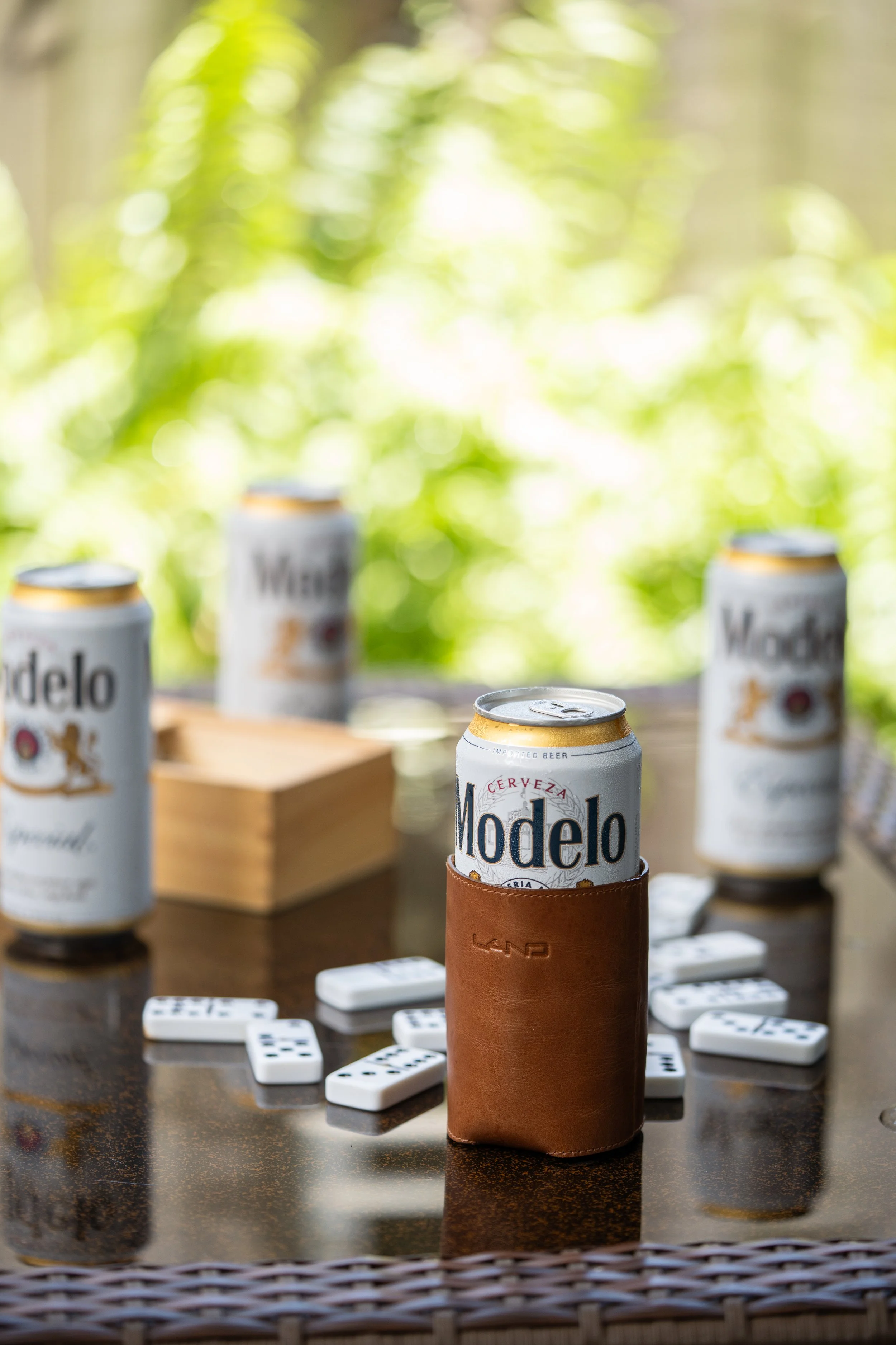 A table with four cans of Modelo beer, some dominoes, and a leather holder on it, with a blurred green background.