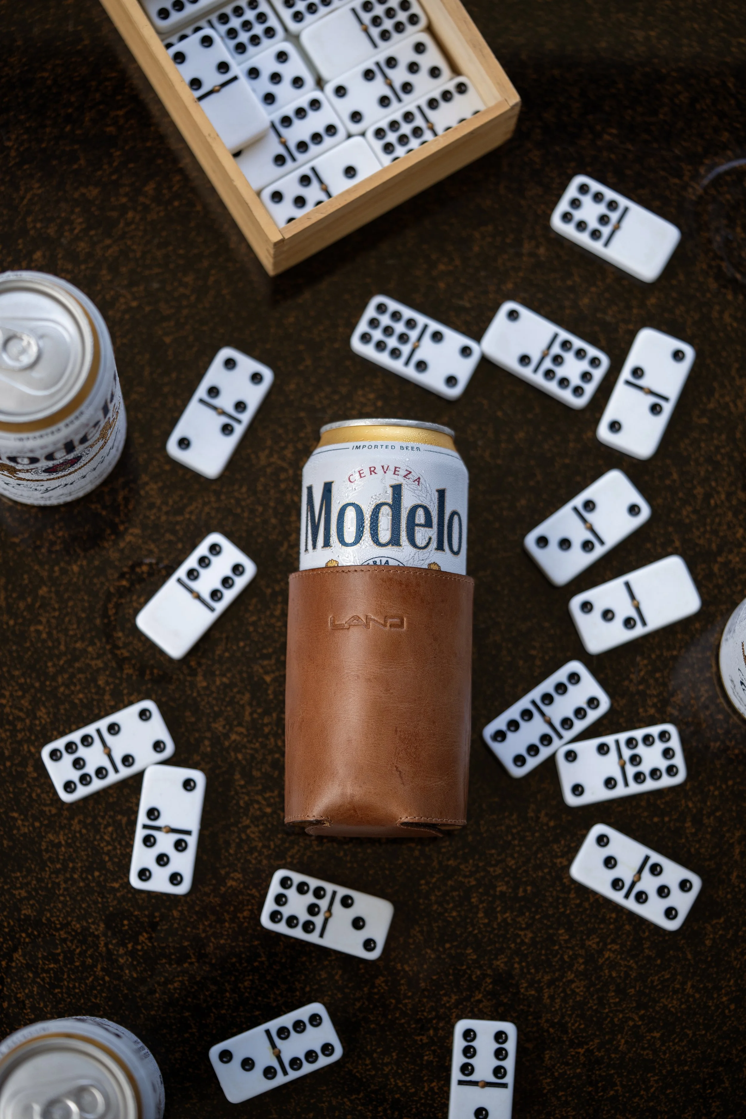 A can of Modelo beer in a leather sleeve surrounded by scattered dominoes and a box of dominoes on a dark surface.
