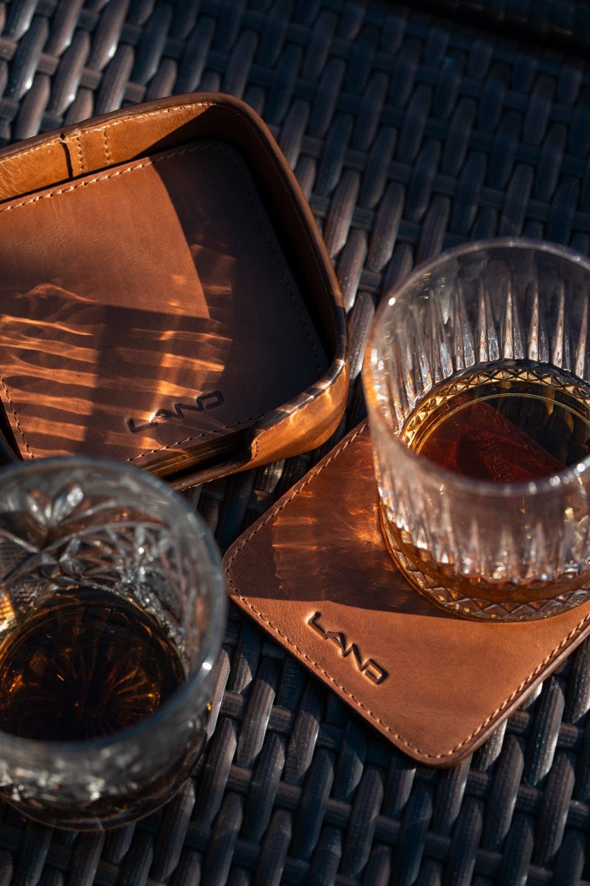 Two glasses of dark liquor on woven dark table, each placed on a leather coaster with the brand name 'LANZ'.