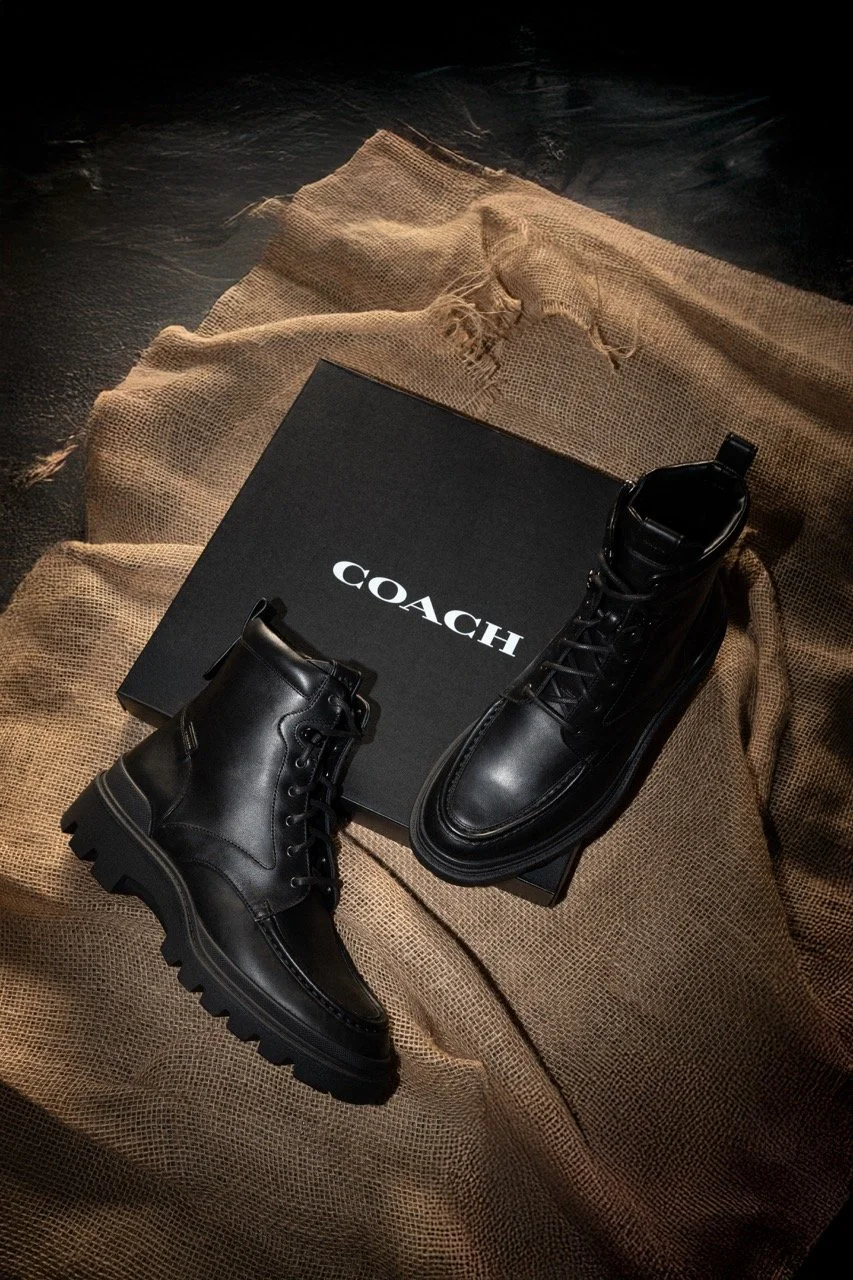 Pair of black leather boots placed on a black COACH shoe box, with a textured burlap fabric background.
